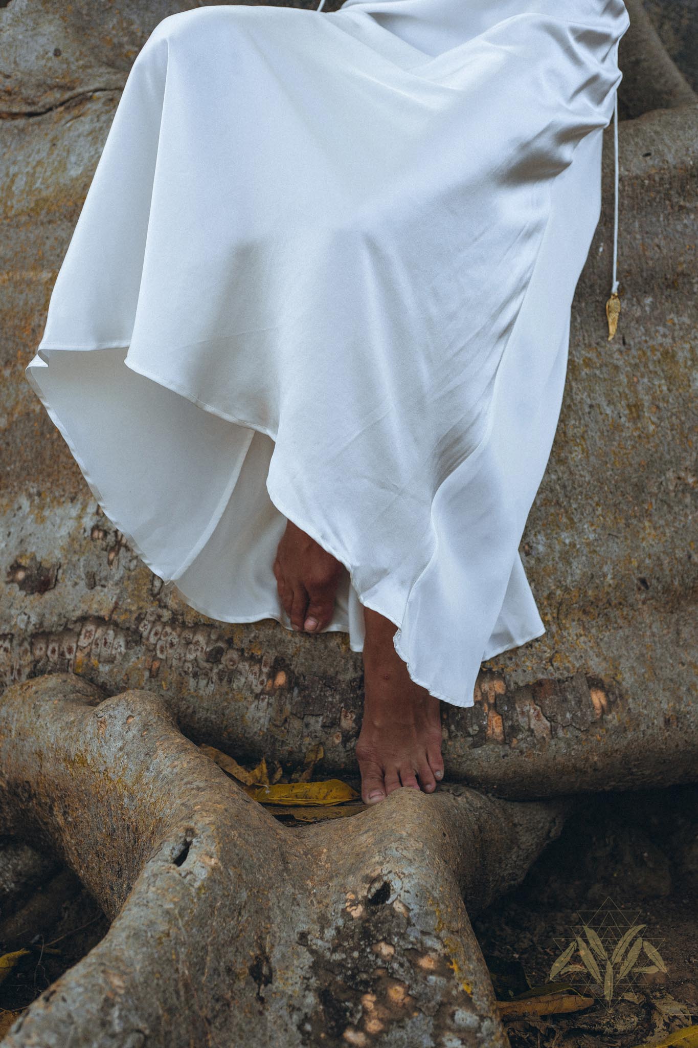 A person wearing the White Peace Silk Simple Wedding Dress by AYA Sacred Wear stands barefoot on large tree roots. The natural silk of the boho dress cascades gracefully, contrasting with the rugged texture of the roots.