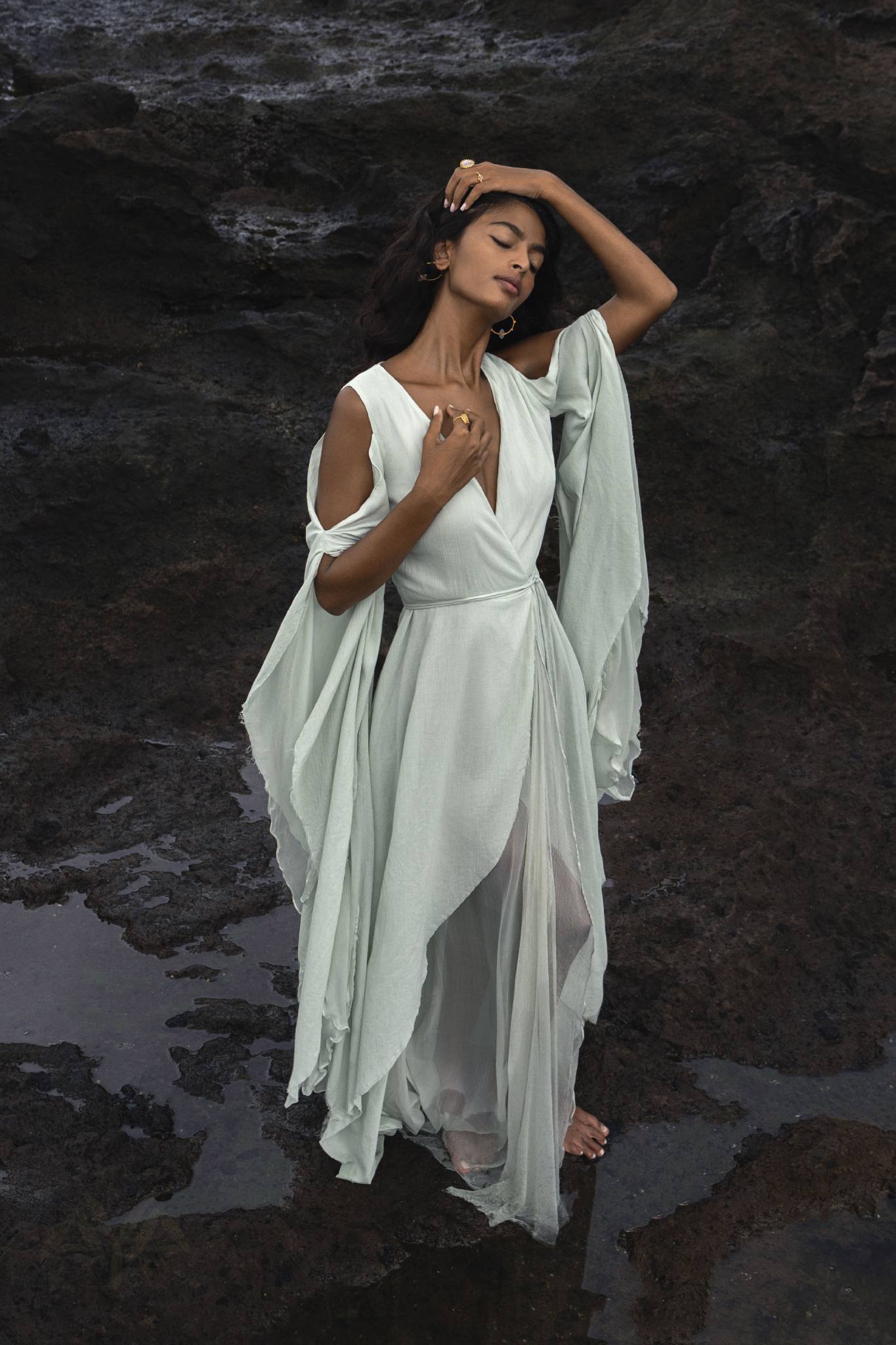 A woman poses gracefully with one hand on her head, eyes closed, wearing an enveloping, sky-blue Makosh Goddess Dress by AYA Sacred Wear. The dress features draped sleeves and flows elegantly against the dark, rocky terrain backdrop.
