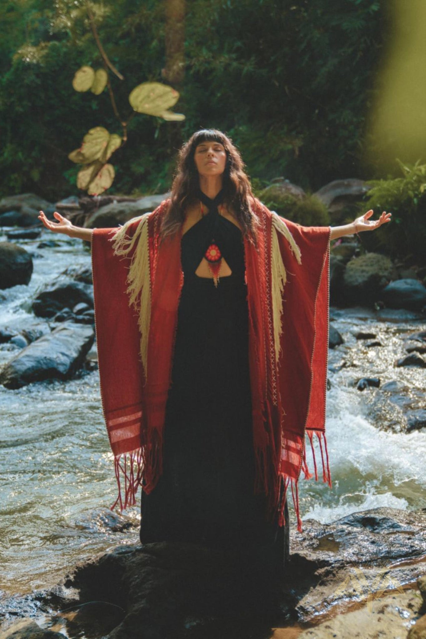 A woman stands on rocks in a stream, arms spread wide and looking upward. She is wearing the Black Harmonia Dress from AYA Sacred Wear, a Gothic sexy, adjustable dress also known as the Fairy of the River Dress. The dress is crafted from organic linen and cotton and is complemented by a red and beige shawl. The surrounding area is lush with greenery as sunlight filters through the trees.