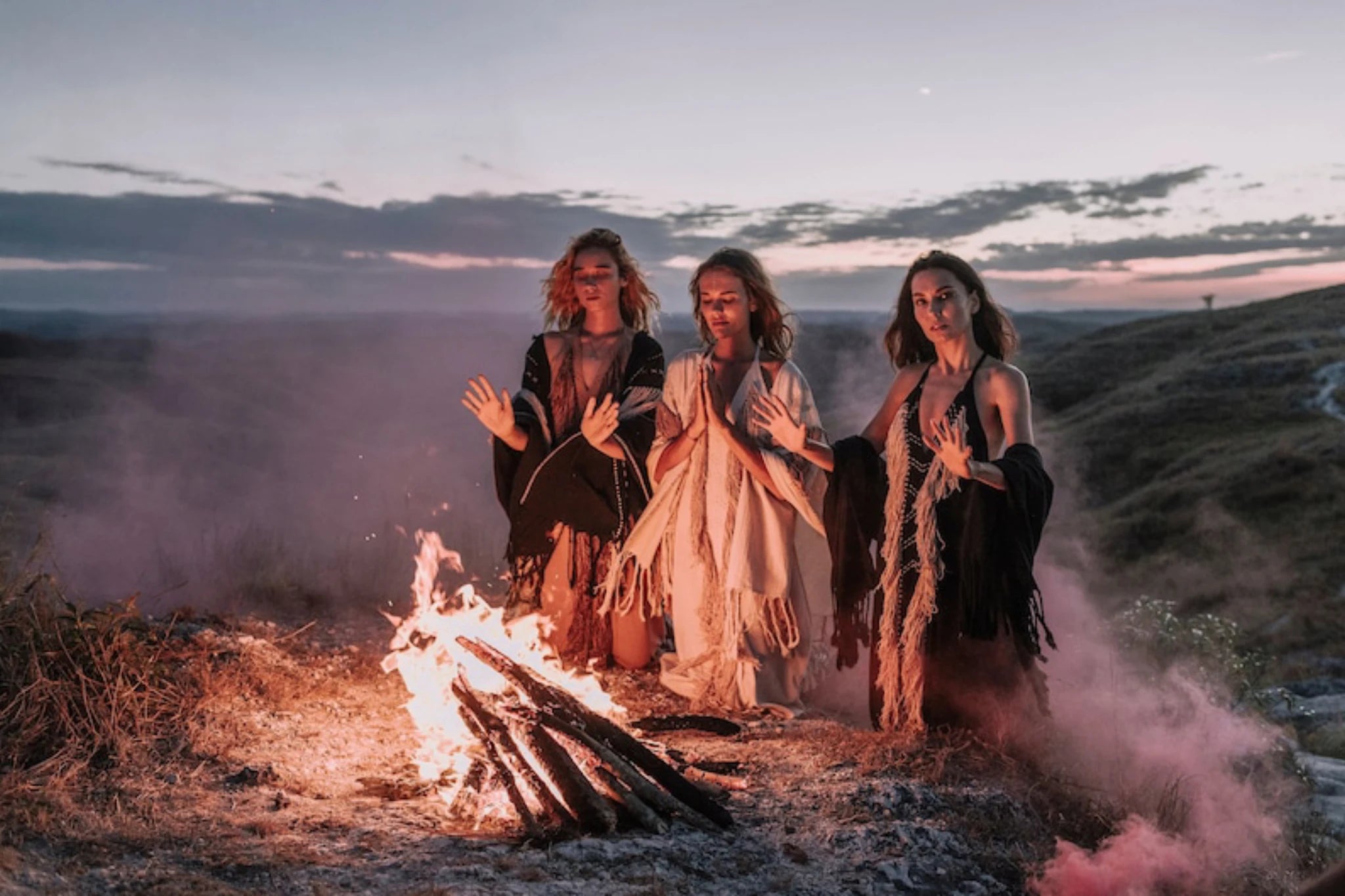 Three women in AYA Sacred Wear’s Black Cotton Long Maxi Dresses with open backs warm their hands by a bonfire at dusk, smoke swirling around them. Their goddess-inspired dresses are crafted from organic cotton.