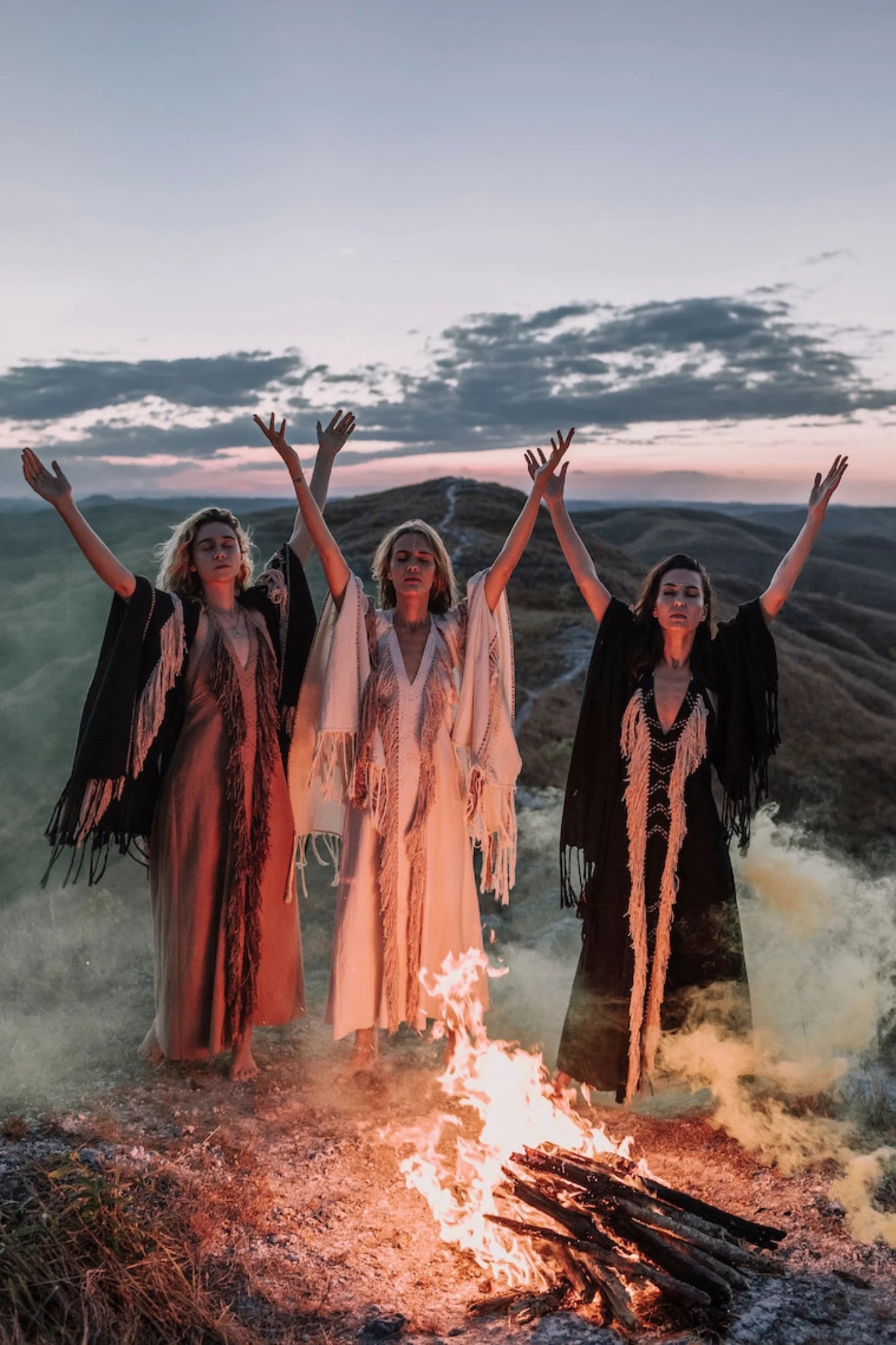 Three women in Black Cotton Long Maxi Dresses by AYA Sacred Wear stand on a hilltop at dusk, arms raised to a campfire. The dramatic sky and drifting smoke create a mystical atmosphere around them.