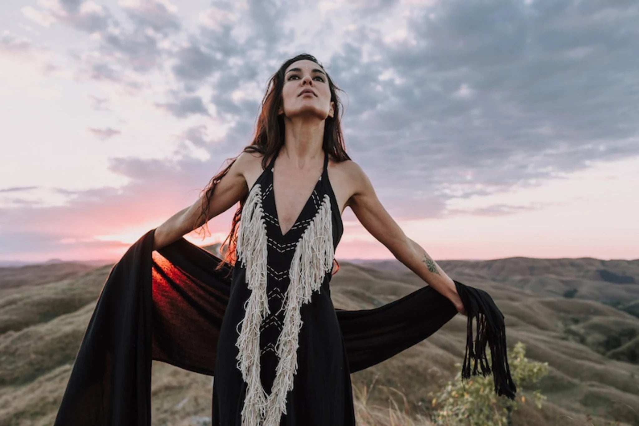A woman in the AYA Sacred Wear Black Cotton Long Maxi Dress with an open back stands outdoors at sunset, holding out her goddess dress. Rolling hills and dramatic clouds fill the background.