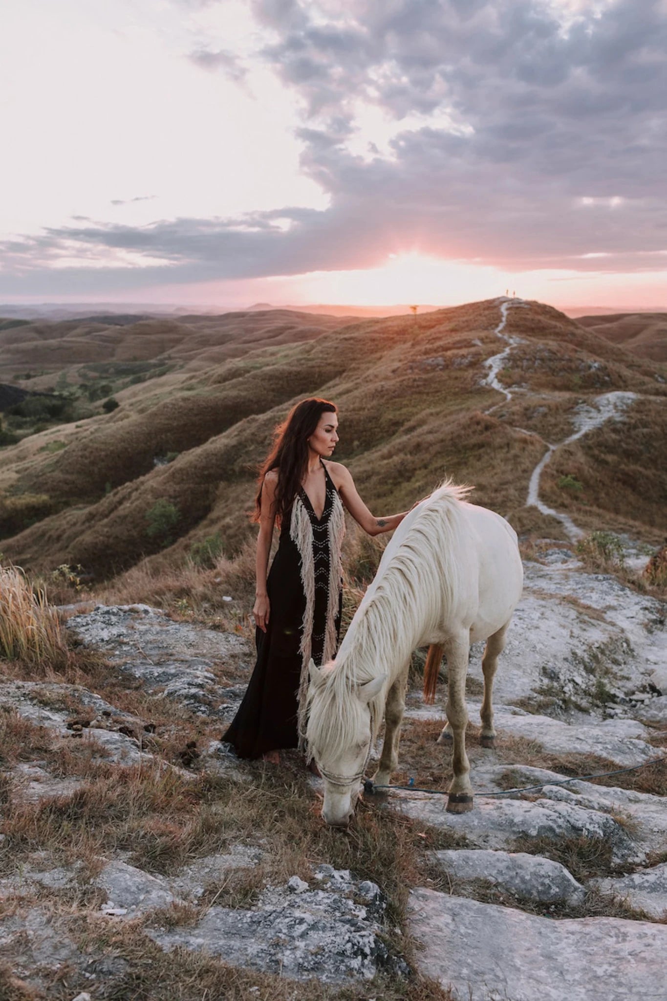 A woman wearing the Black Cotton Long Maxi Dress • Open Back Goddess Dress by AYA Sacred Wear stands on rocky terrain at sunset, gently touching a grazing white horse as rolling hills and a winding path extend beneath a dramatic sky.