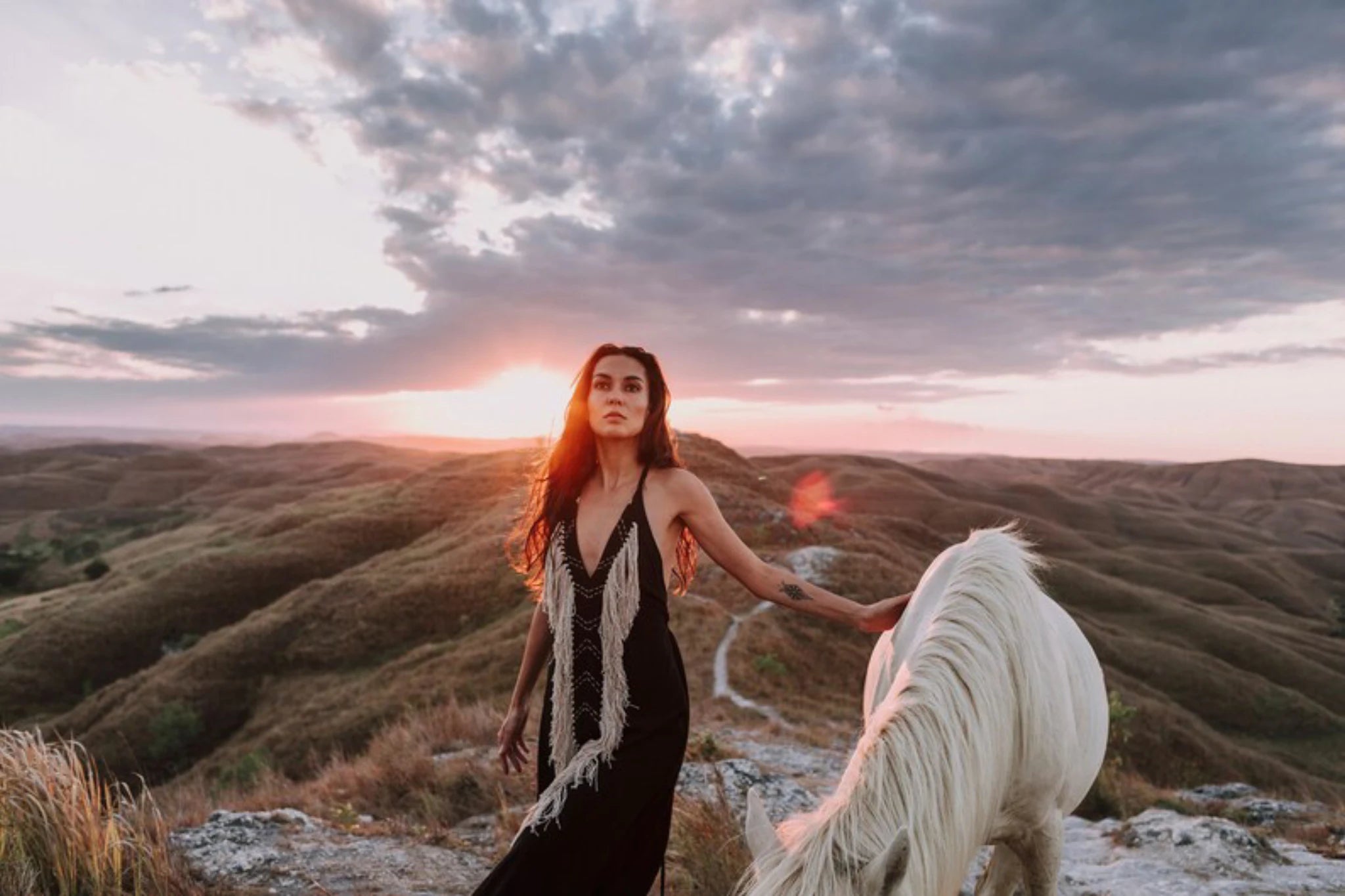 A woman in a Black Cotton Long Maxi Dress by AYA Sacred Wear stands on a hill at sunset, gently touching a white horse, with rolling hills and dramatic clouds in the background. The Open Back Goddess Dress is crafted from organic fabric and eco-friendly dyes.