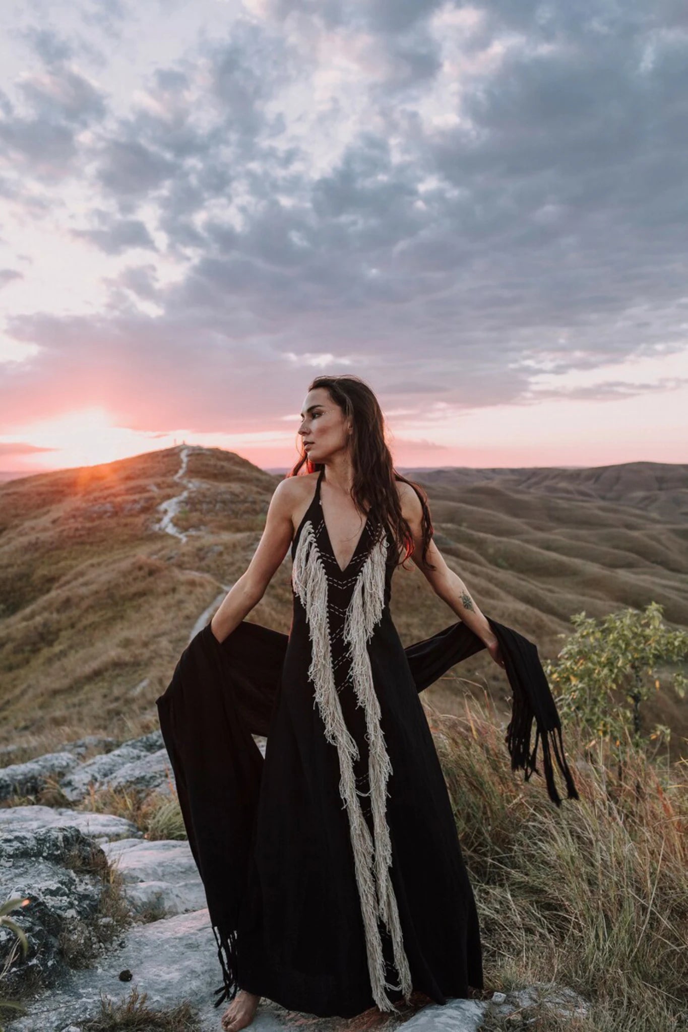 A woman in the Black Cotton Long Maxi Dress by AYA Sacred Wear stands barefoot on rocky ground at sunset with rolling hills and a cloudy sky behind her, gazing to the side and holding out the sides of her open-back goddess dress.