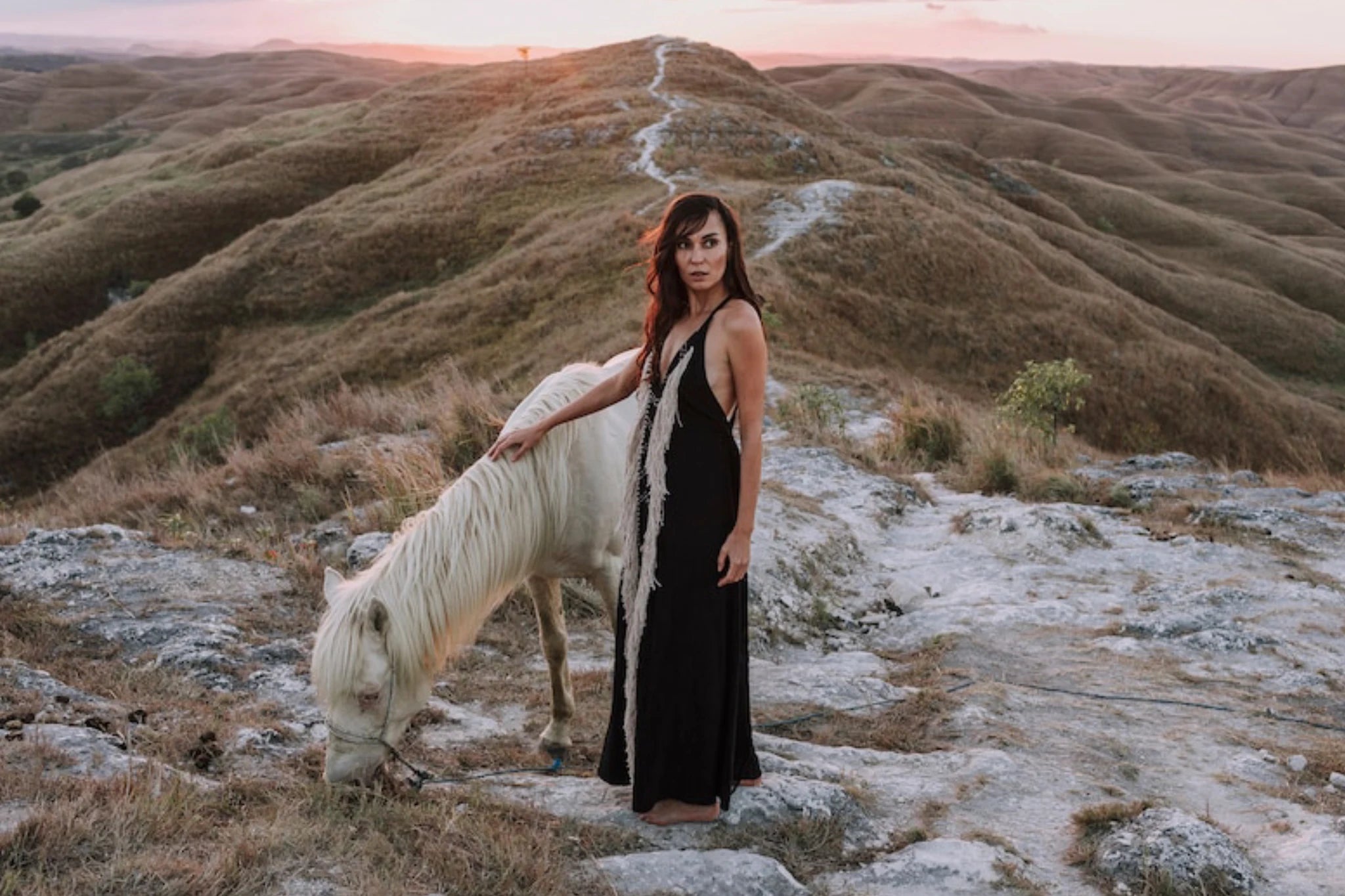 A woman wearing the Black Cotton Long Maxi Dress by AYA Sacred Wear stands on rocky ground at sunset, gently touching a grazing white horse as rolling brown hills and a winding path stretch into the background.