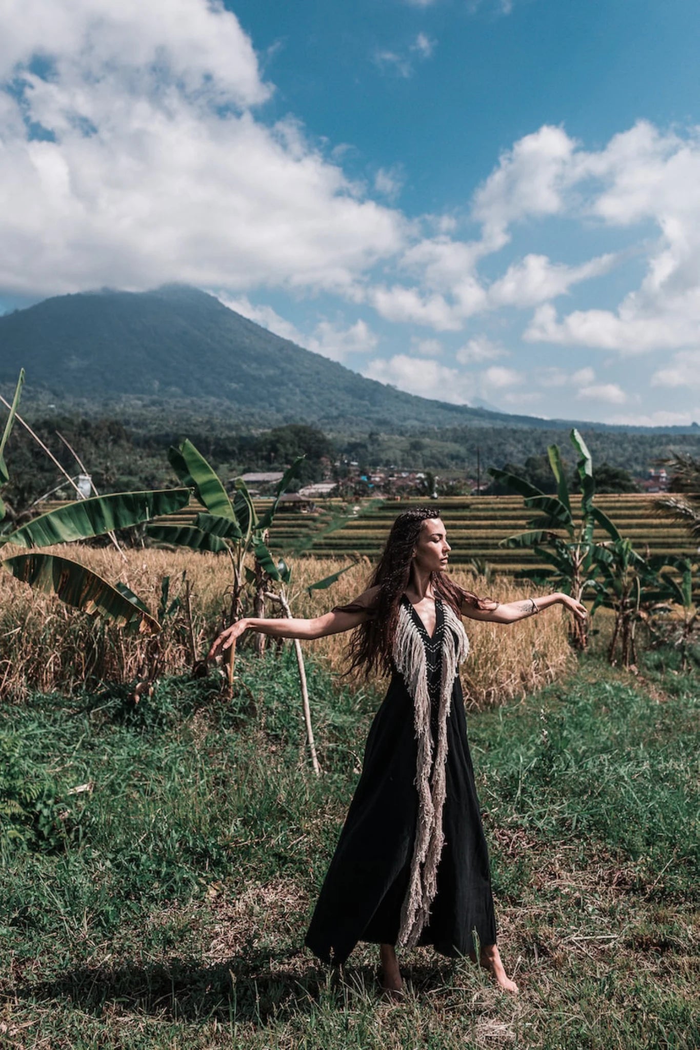 A woman wearing the AYA Sacred Wear Black Cotton Long Maxi Dress • Open Back Goddess Dress poses with outstretched arms in a lush field of banana plants and golden crops, with mountains and a cloudy blue sky in the background.