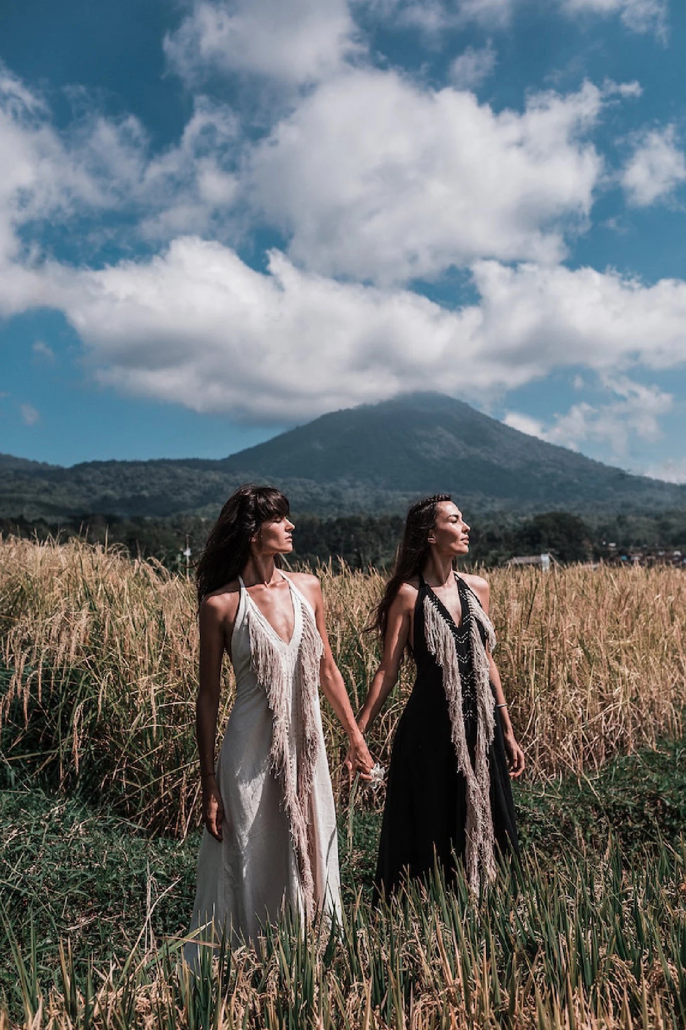 Two women wearing AYA Sacred Wear's Black Cotton Long Maxi Dress with open backs stand hand in hand in a grassy field, with mountains and a cloudy sky behind them.