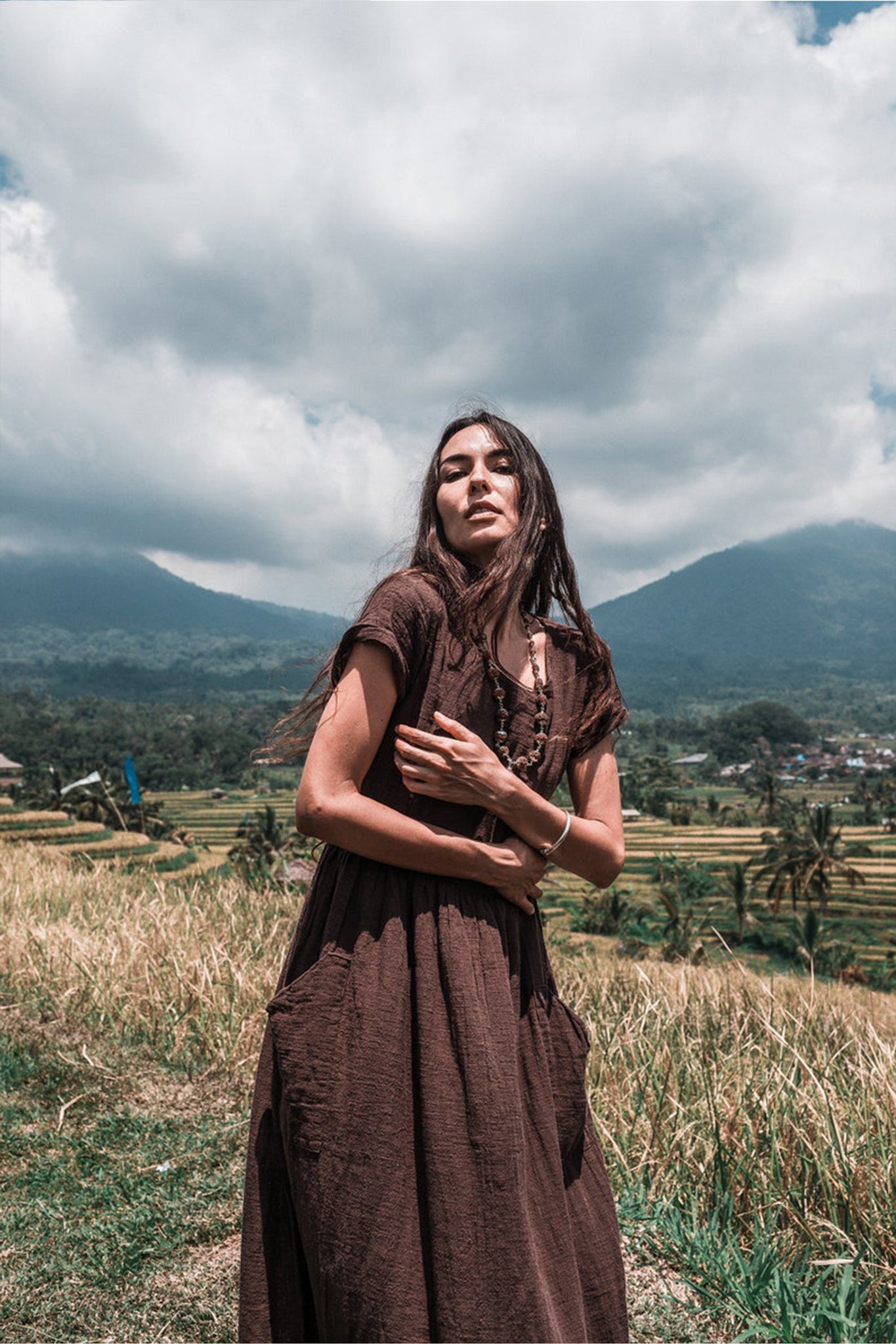 In a serene grassy field with mountains and cloudy skies as the backdrop, a woman showcases her style in a Bohemian Boho Dress for Women by AYA Sacred Wear. Made from handwoven raw cotton, this dress embodies her dedication to sustainable ethical fashion. Her long hair gently sways in the breeze, harmonizing perfectly with the tranquil natural environment.
