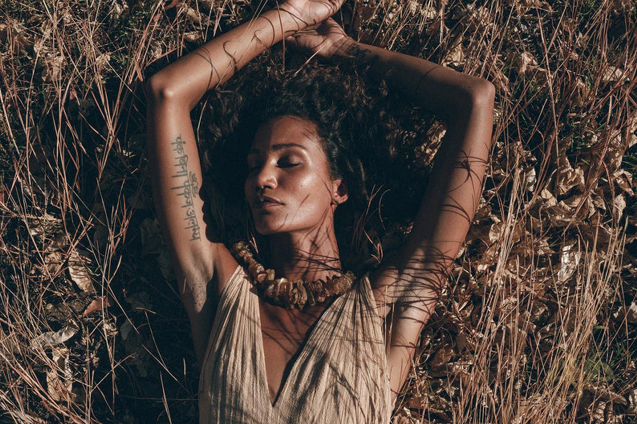 A woman rests in a field of dry grass, her arms stretched above her head. She is wearing a Beige Sandal Organic Dress from AYA Sacred Wear, featuring handmade braided straps, and accessorizes with a necklace adorned with large beads. Her eyes are closed as the shadows of branches drape across her face and arms.