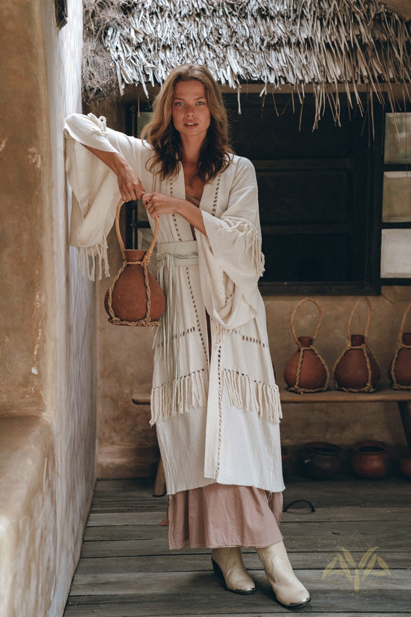 In a rustic setting over a wooden floor, a woman stands under the thatched roof wearing AYA Sacred Wear's Boho Kimono Cardigan, featuring handwoven natural cotton with fringe detailing. Holding a large jug amidst similar jugs, her light brown hair and white boots perfectly complement her look.