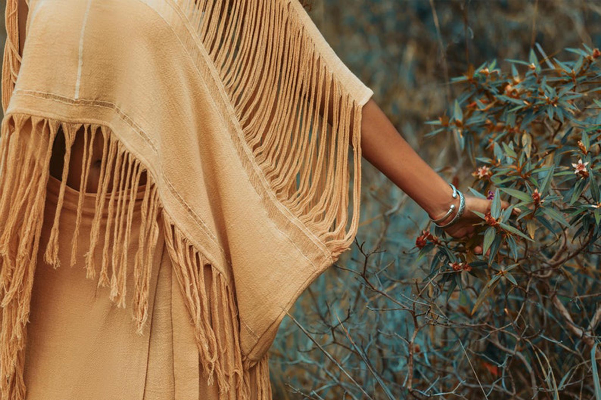 A person wearing the AYA Sacred Wear Boho Top, a light brown hand-loomed cotton blouse, stands in a natural setting, gently touching the leaves of a plant. Their arm is adorned with silver bracelets, and the scene evokes a sense of tranquility and connection with nature. The hand-loomed cotton adds an earthy, sustainable touch.