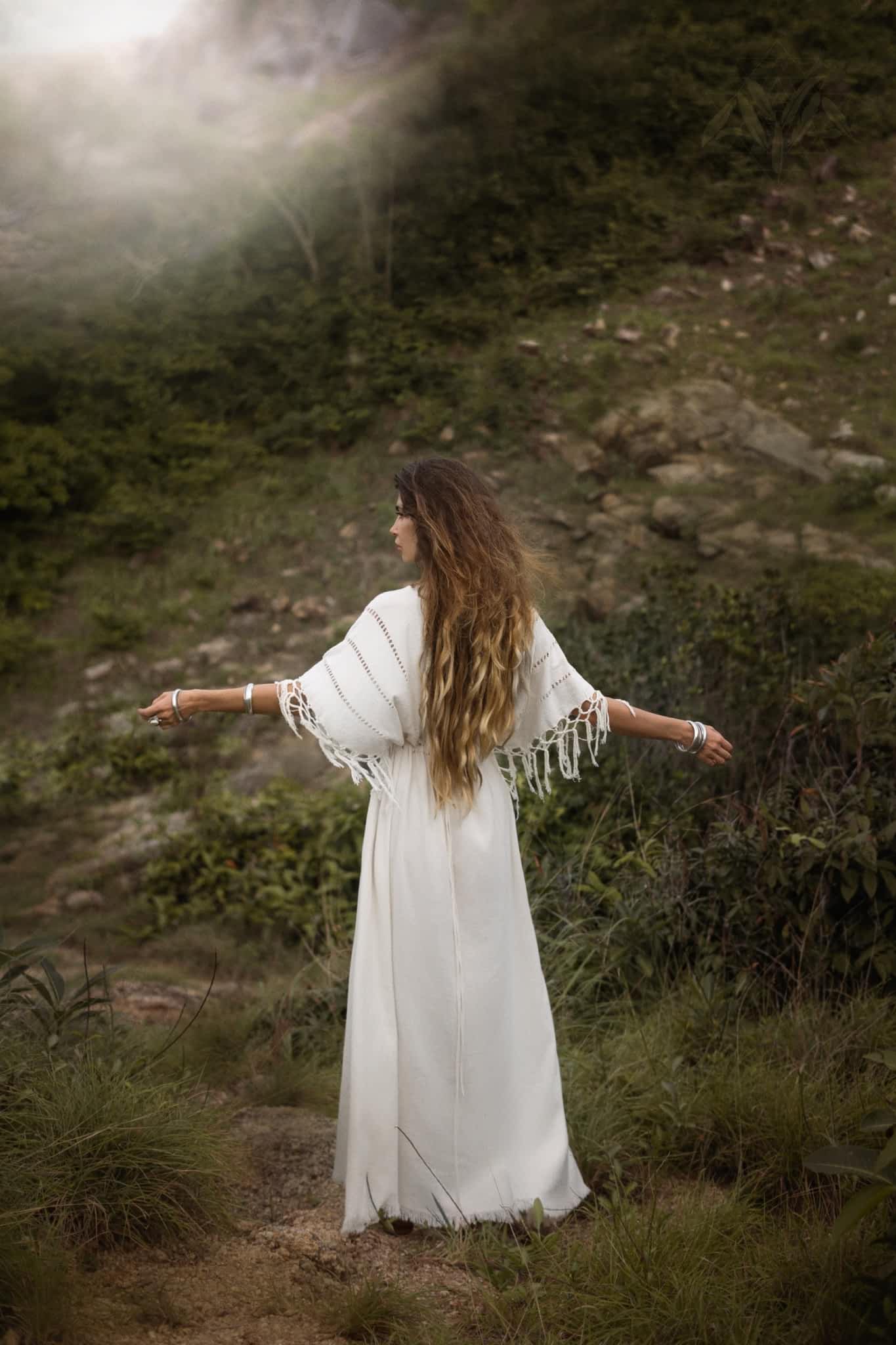 A woman with long hair wears an AYA Sacred Wear Off-White Boho Goddess Dress, arms outstretched in a grassy, rocky landscape. Facing away, she's enveloped by greenery and a misty, serene atmosphere.