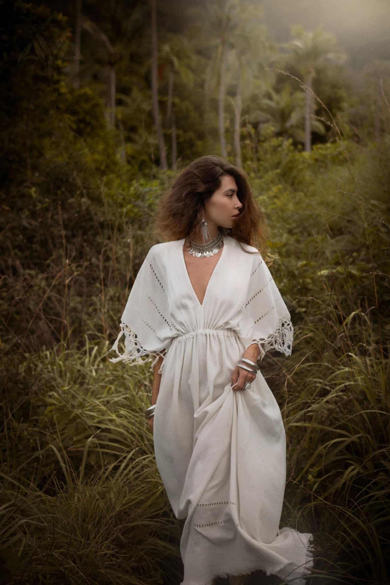In a lush green forest, a woman stands wearing an AYA Sacred Wear Off-White Boho Dress adorned with traditional handmade needlework. Her long brown hair and silver jewelry accentuate the misty, serene atmosphere of tall trees surrounding her.