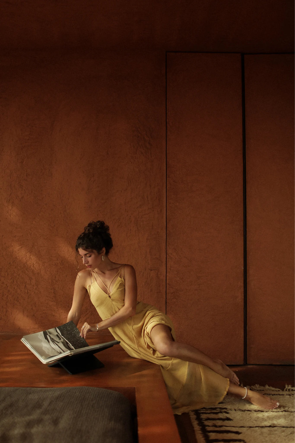A woman in the Powder Yellow Padma Dress by AYA Sacred Wear sits on a patterned rug, leaning against wood, reading a large book in a warmly lit room with earthy brown walls.