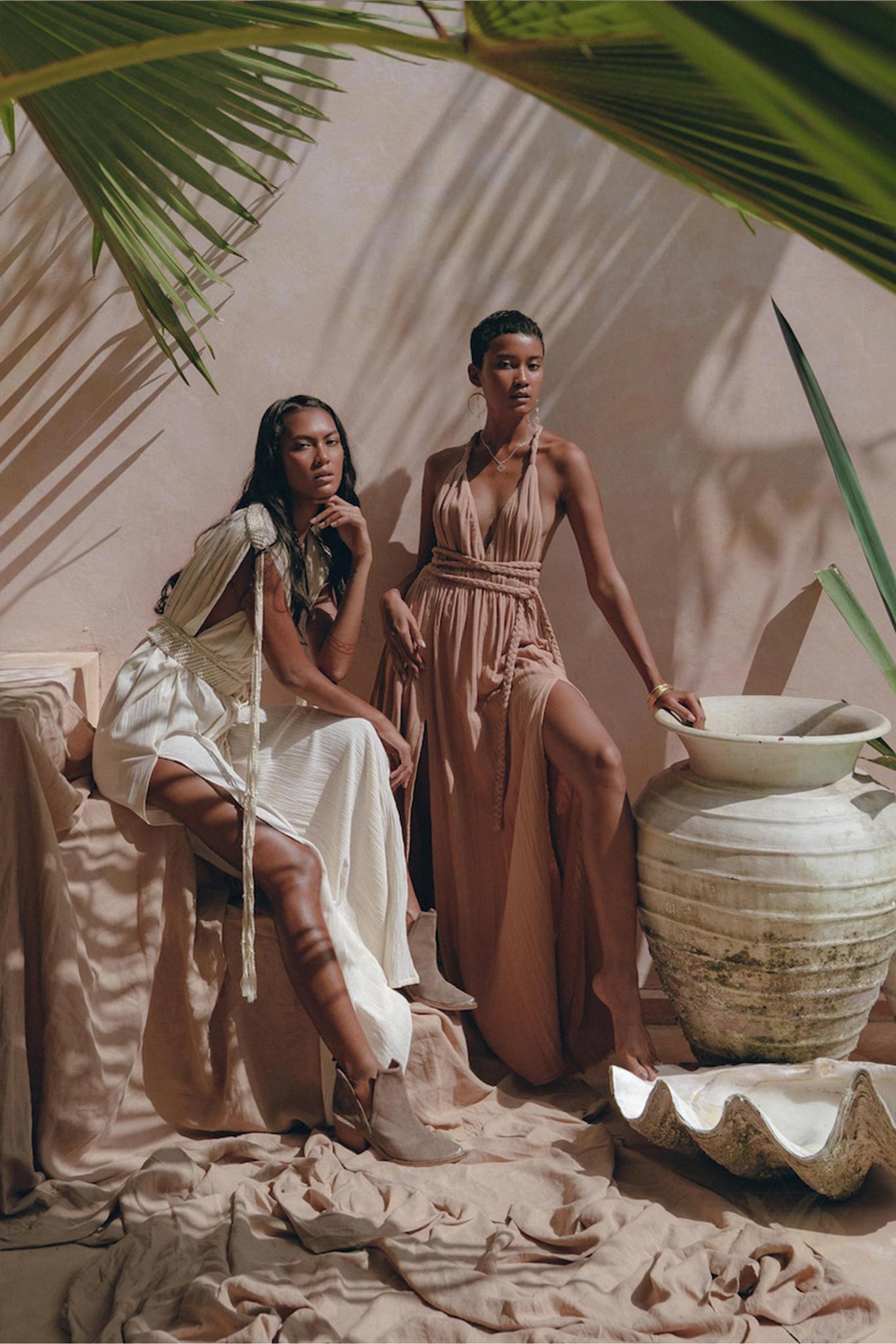 Two women pose in an earthy, desert-inspired setting. One is wearing a flowing boho bridesmaid dress paired with boots while sitting on a draped surface. The other stands beside a large ceramic vase, showcasing the Dusty Pink Greek Goddess Dress from AYA Sacred Wear. Large green leaves frame the scene beautifully.