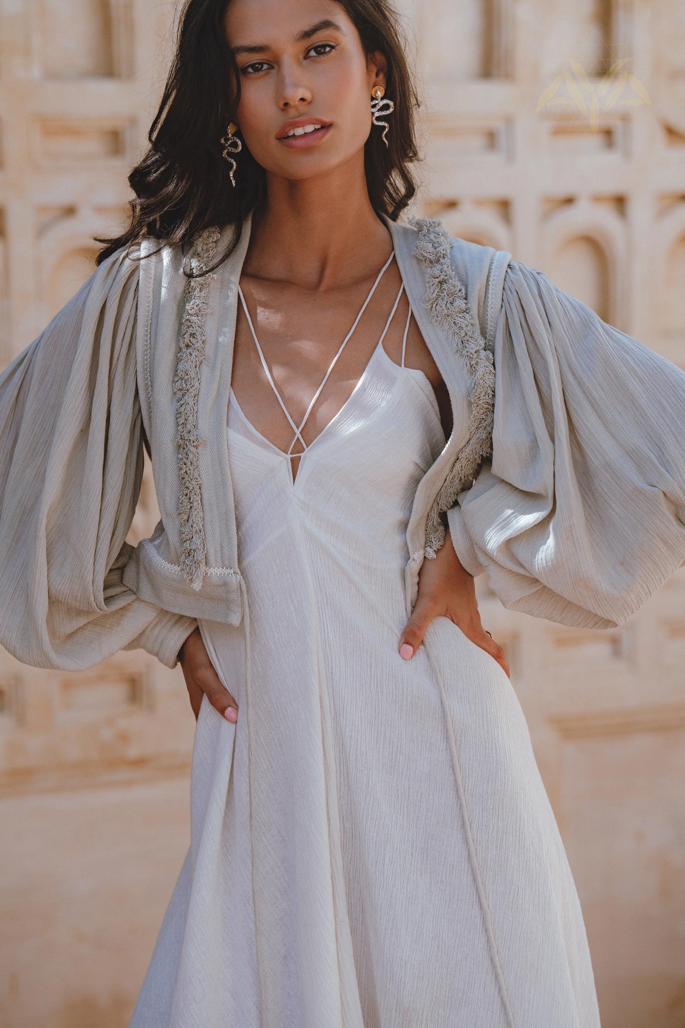 A model wearing a dusty blue puff sleeves top with organic fabric, featuring boho tassels. The top is styled over a white dress, showcasing a relaxed yet elegant look against a warm, textured background. Perfect for tropical settings.