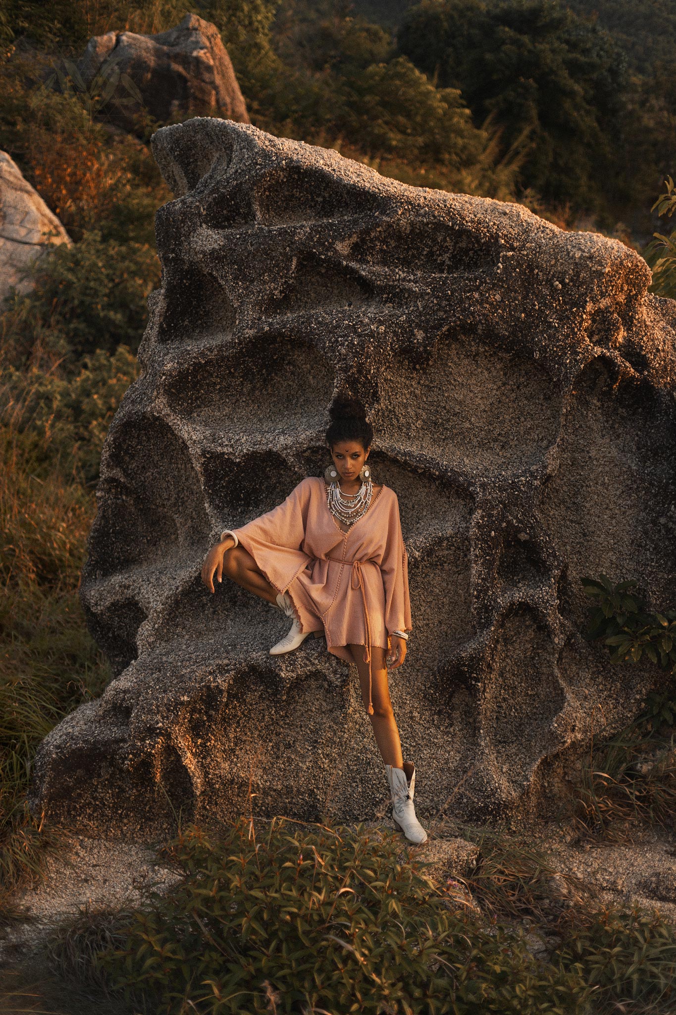 A model poses dramatically on a textured rock formation, wearing a dusty pink handmade knotted jumpsuit. The outfit has a relaxed fit with billowy sleeves, accentuated by a decorative tie at the waist. She pairs the look with stylish white boots, and layered statement jewelry adorning her neck. The backdrop features lush greenery under warm, golden lighting, creating an ethereal atmosphere. This ensemble reflects the aesthetics of the Dusty Pink Handmade Knotted Jumpsuit, a unique piece perfect for various