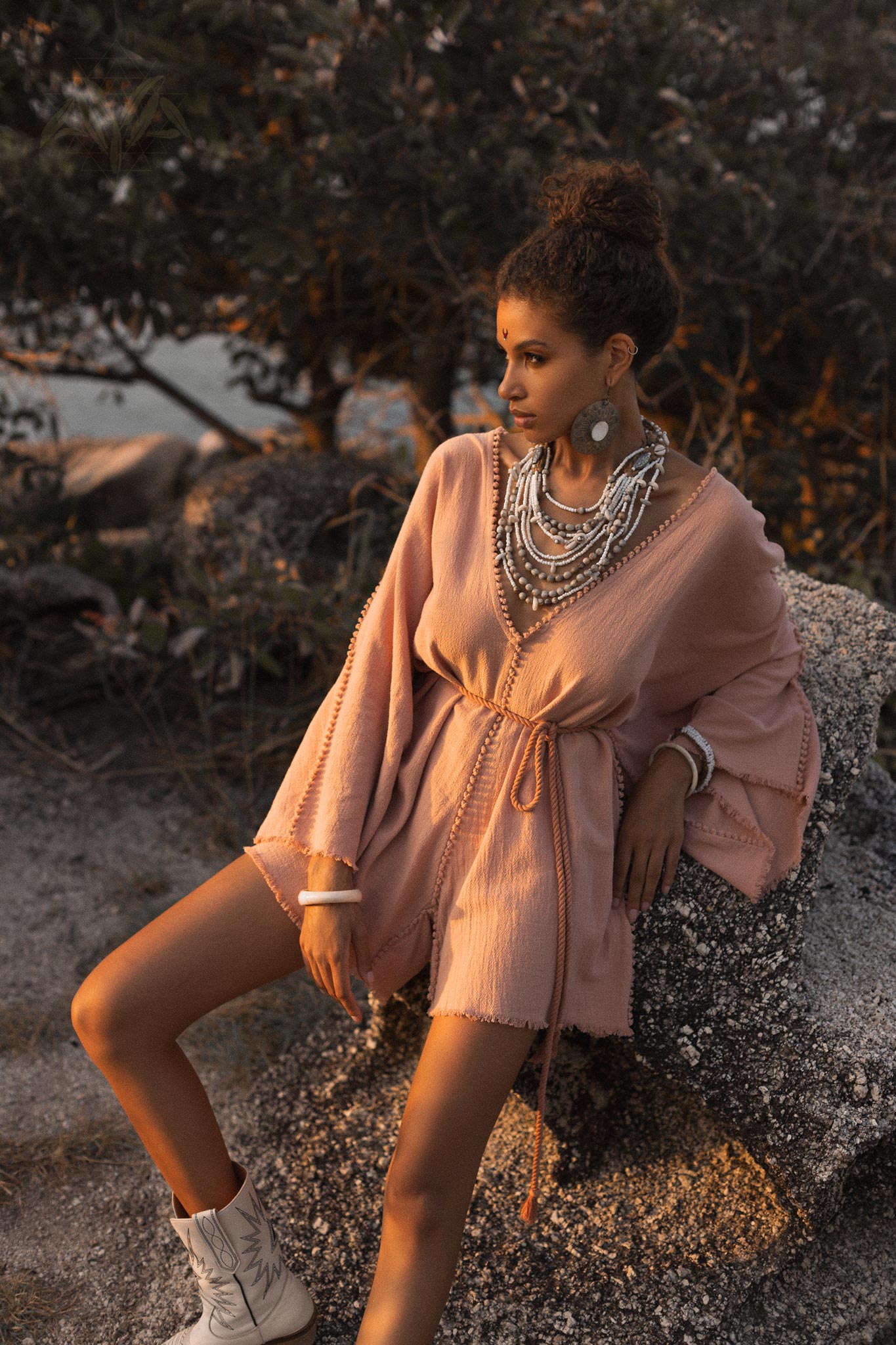 A model poses gracefully in a dusty pink handmade knotted jumpsuit romper dress, adorned with layered necklaces. The outfit features loose sleeves and a tied waist, set against a natural background of rocks and greenery, with warm light illuminating the scene, evoking a relaxed yet stylish vibe. This elegant attire is ideal for bridesmaids, capturing a goddess-like aesthetic by Aya Sacred Wear.