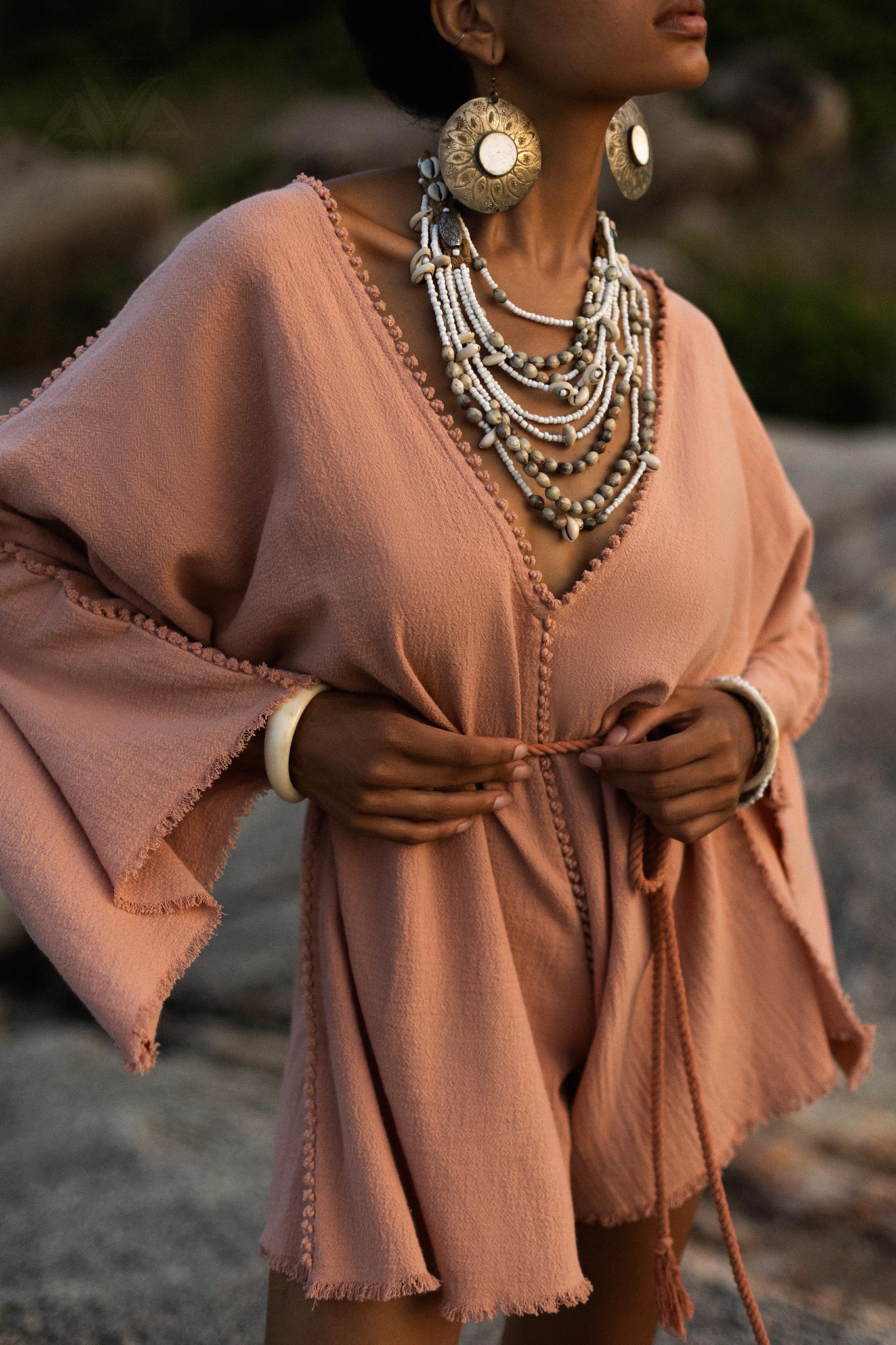 A model showcases a dusty pink handmade knotted jumpsuit, featuring a relaxed fit with flared sleeves and decorative trim. The outfit is complemented by layered, intricate beaded jewelry and large statement earrings, creating an elegant bohemian look. The backdrop features natural elements, enhancing the organic vibe of the ensemble. This stylish piece is perfect for various occasions, merging comfort with chic aesthetics.