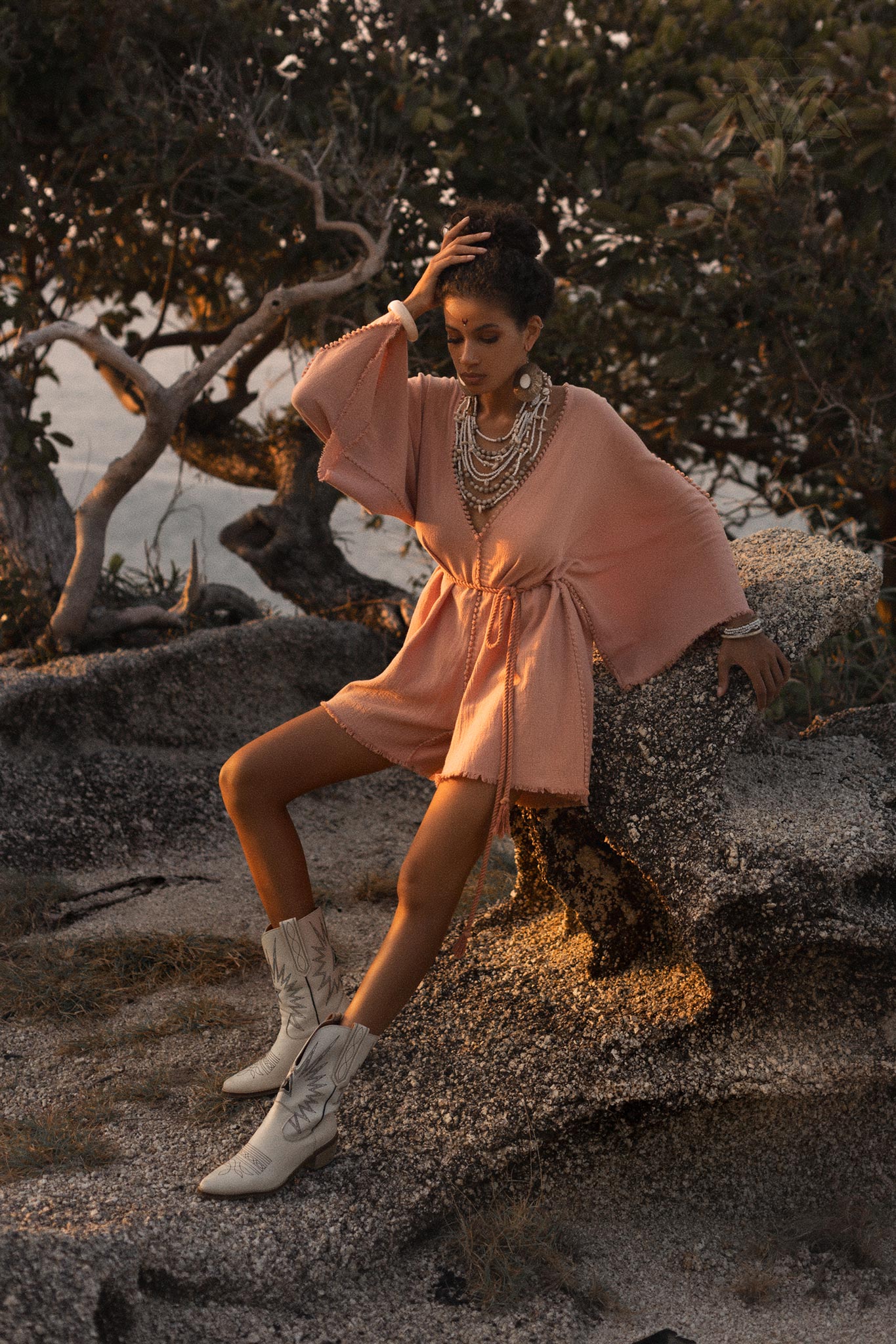 A model poses in a dusty pink handmade knotted jumpsuit, featuring wide sleeves and a cinched waist. She pairs the outfit with stylish white cowboy boots, showcasing layered necklaces that add an elegant touch. The backdrop consists of natural landscapes, with warm lighting enhancing the overall aesthetic. This attire is perfect for a romantic occasion or a bohemian-themed event, showcasing the beauty of organic fashion choices.