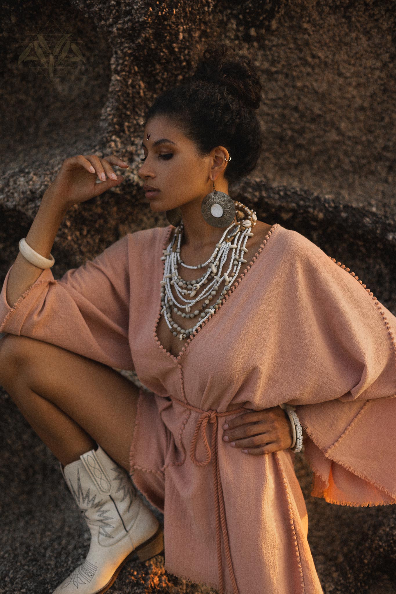 A woman poses elegantly in a dusty pink handmade knotted jumpsuit, showcasing its organic design. She has layered intricate necklaces and wears distinctive earrings, enhancing her sophisticated look. The setting features textured earthy tones, complementing her attire. This outfit is perfect for occasions like bridesmaid events or modern gatherings.