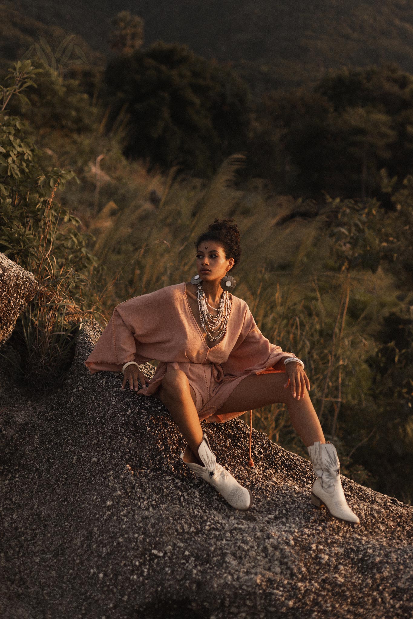 A model poses on a rocky surface, wearing a dusty pink handmade knotted jumpsuit. The outfit features a loose fit with wide sleeves and a tie at the waist. She accessorizes with multiple layers of necklaces and large earrings, complemented by white cowboy boots. The background showcases lush greenery and mountains, illuminated by golden hour light, creating a serene and stylish atmosphere. This look embodies a blend of elegance and bohemian style, perfect for occasions like bridesmaid events or casual outin