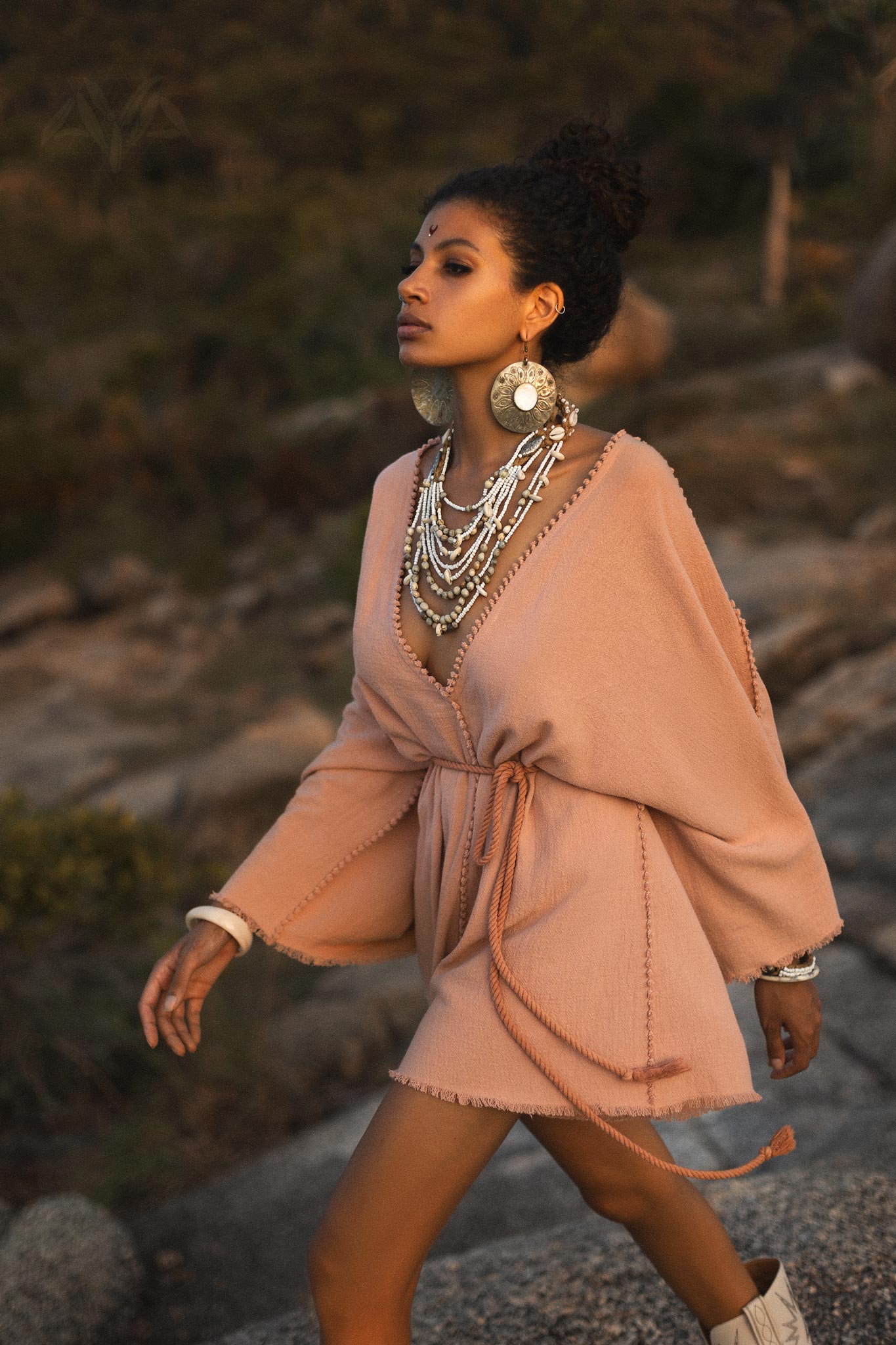 A model walks gracefully along a rocky outdoor setting, showcasing a dusty pink, handmade knotted jumpsuit. The outfit features a V-neckline, wide sleeves, and a belted waist, highlighting its organic fabric. The model accessorizes with large, silver statement earrings and multiple layers of intricate necklaces, enhancing the bohemian style. The warm tones of the setting sun illuminate the natural backdrop, creating an enchanting atmosphere suitable for showcasing the unique design.