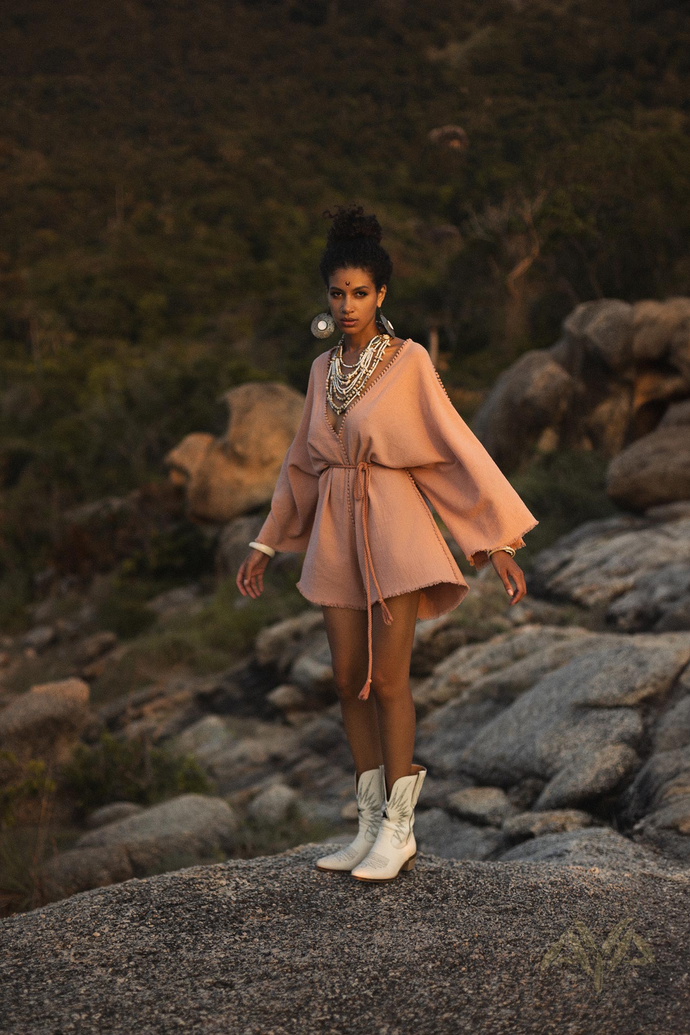 A model stands confidently on a rocky surface, wearing a dusty pink handmade knotted jumpsuit that resembles a mini romper dress. The outfit features a deep V neckline, wide sleeves, and a tie at the waist, accentuating her silhouette. She accessorizes with multiple layers of statement necklaces and oversized earrings. Complementing her look, she wears white cowboy boots, and the natural landscape behind her includes lush greenery and rugged rocks, creating a serene outdoor setting. This design is part of t