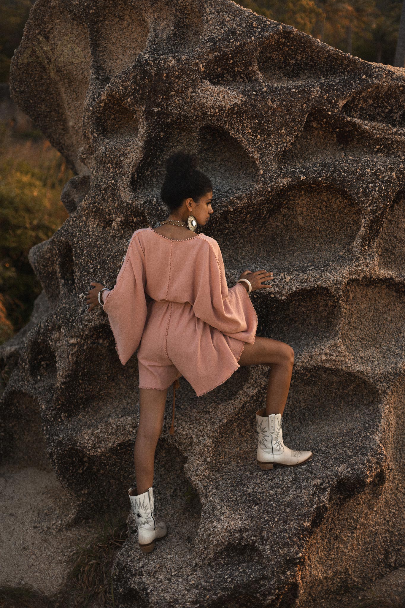 A model wearing a dusty pink, handmade, knotted mini jumpsuit is climbing a rocky surface. The jumpsuit features flowing sleeves and a comfortable fit. She pairs the outfit with white cowboy boots. The background showcases textured rocks and warm, natural lighting, enhancing the bohemian aesthetic of the scene. This look is perfect for casual styling or as a bridesmaid option by Aya Sacred Wear.