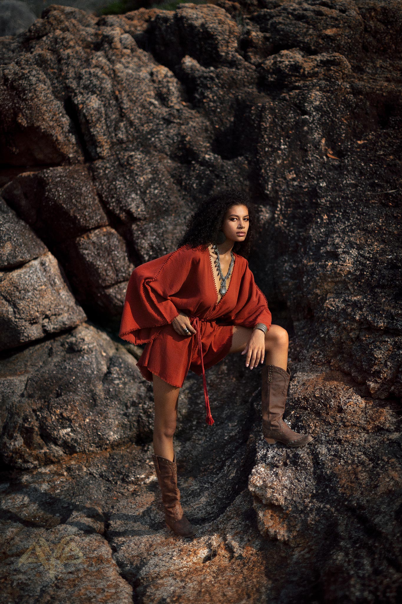 A woman poses confidently on rocky terrain, wearing a red handmade knotted jumpsuit. The outfit features a relaxed fit and a stylish belt. She complements the look with knee-high brown boots and layered jewelry. The natural surroundings enhance the organic vibe of the outfit. Ideal representation of a red mini organic jumpsuit romper dress for women.