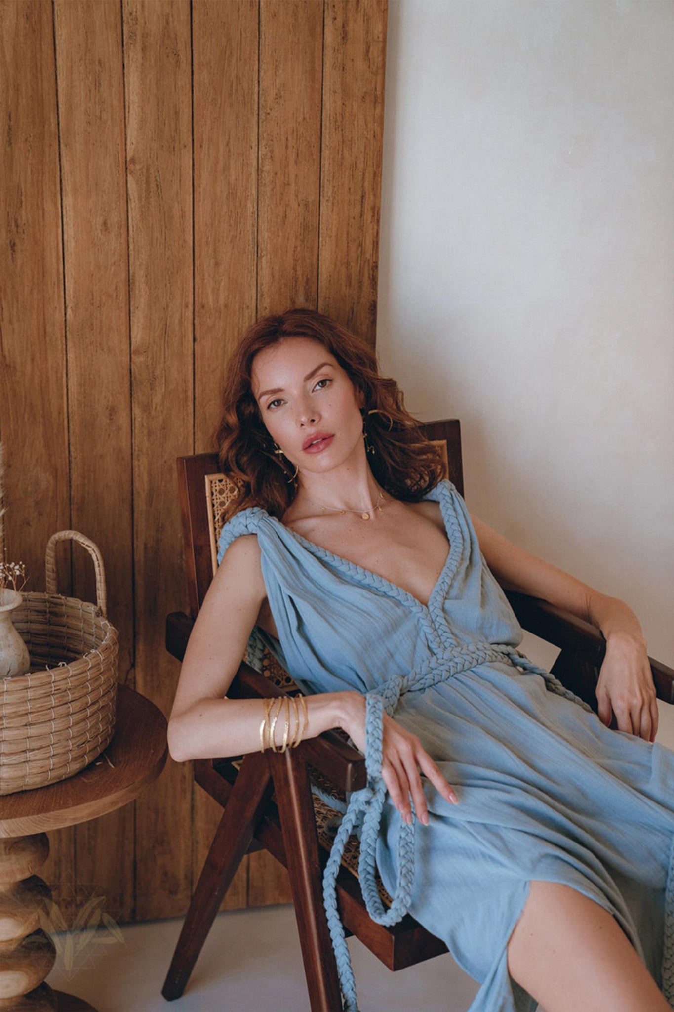 A woman with wavy, reddish-brown hair is lounging in a wooden chair. She is wearing an environmentally friendly Light Blue Nomad Spirit Dress from AYA Sacred Wear, featuring a sleeveless design and subtle jewelry. A woven basket on the side table complements the wooden wall backdrop.