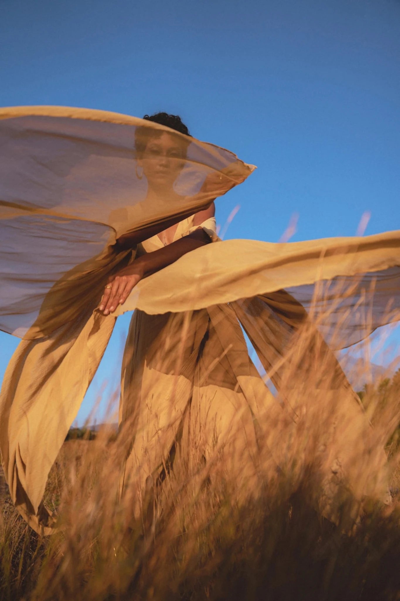 A person stands in tall golden grass, holding the flowing beige fabric of an Ochre Makosh Goddess Dress • Fairy Wings Dress • Light Cotton Formal Dress by AYA Sacred Wear, billowing in the wind under a clear blue sky as sunlight casts warm tones.