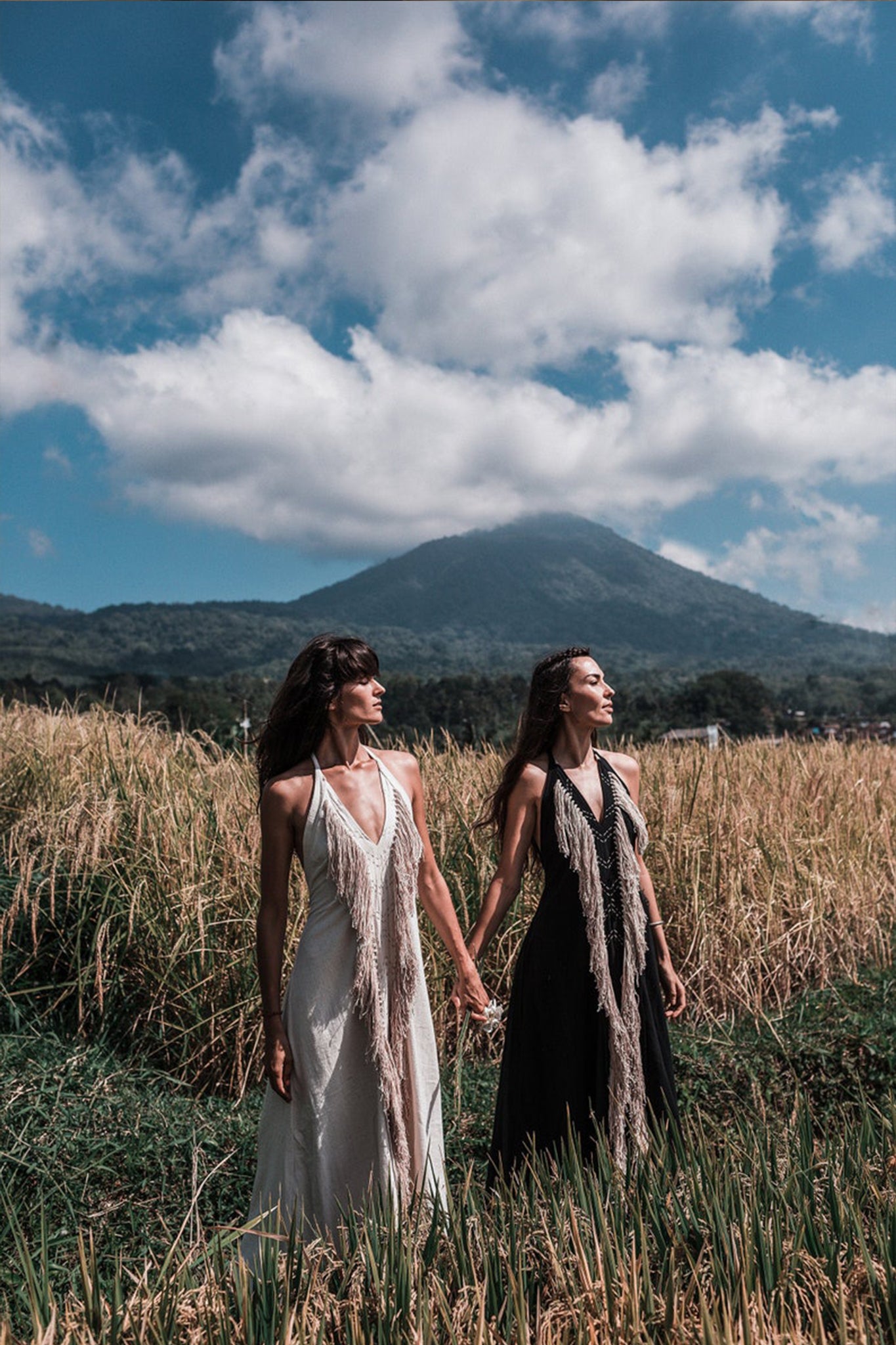 In a lush field, two women stand hand in hand wearing "AYA Sacred Wear" Off-White Boho Tribal Bohemian Dresses. They gaze into the distance, with a majestic mountain rising in the background under a partly cloudy sky. The serene and picturesque scene is beautifully accented by the intricately handmade embroidery on their organic fabric gowns.
