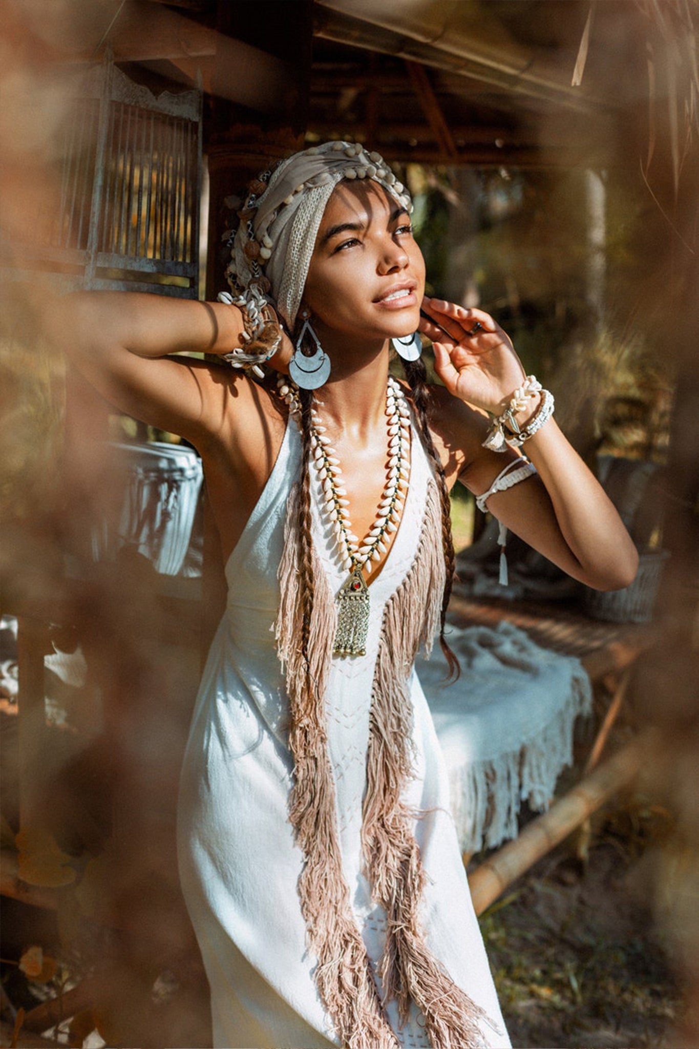 A woman in an off-white Bohemian dress from AYA Sacred Wear stands in a rustic outdoor setting, the entirely handmade tribal design featuring intricate embroidery. She is adorned with layered necklaces and bracelets as dappled sunlight filters through the leaves, creating a serene and dreamy atmosphere.