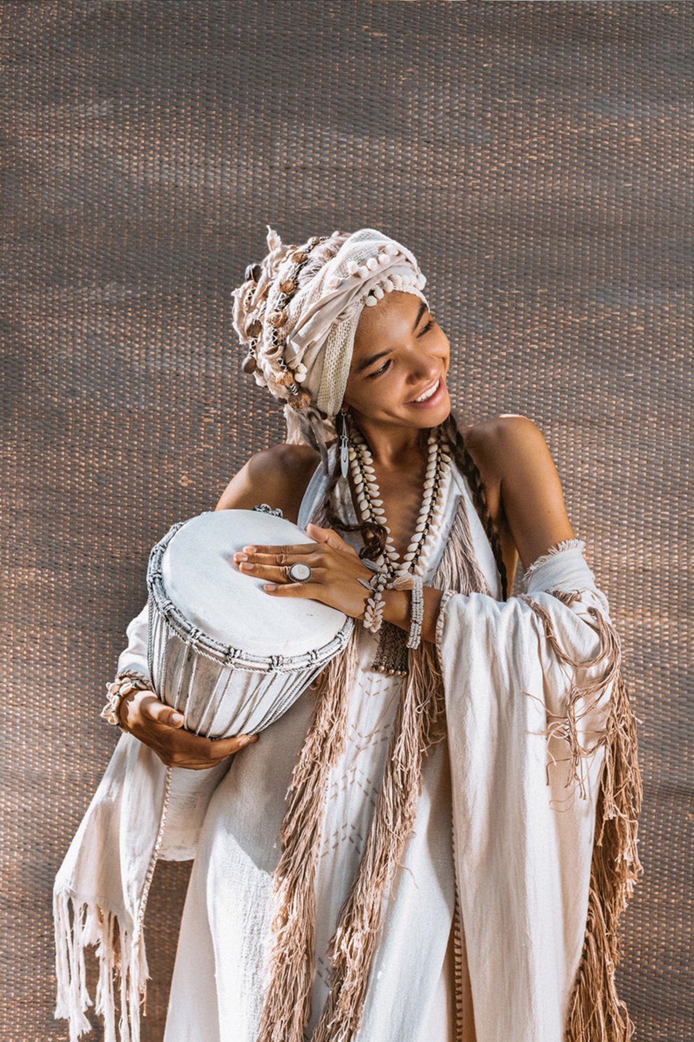 Joyfully playing a traditional hand drum, a person is dressed in the Off-White Boho Tribal Bohemian Dress by AYA Sacred Wear. They wear a headwrap and necklaces, smiling vibrantly against a textured brown background that evokes hand-made embroidery.