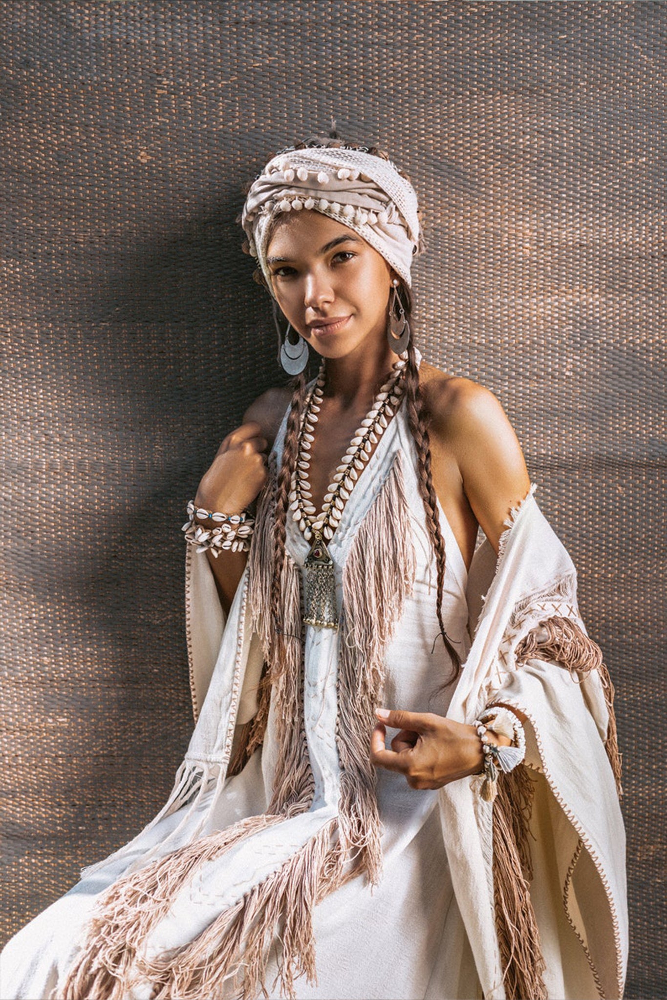 A woman is seated, adorned in AYA Sacred Wear's Off-White Boho Tribal Bohemian Dress, entirely handmade from organic fabric. This exquisite white dress showcases fringed details and comes with a coordinated headscarf. She complements her look with large earrings and necklaces, set against a textured brown backdrop.