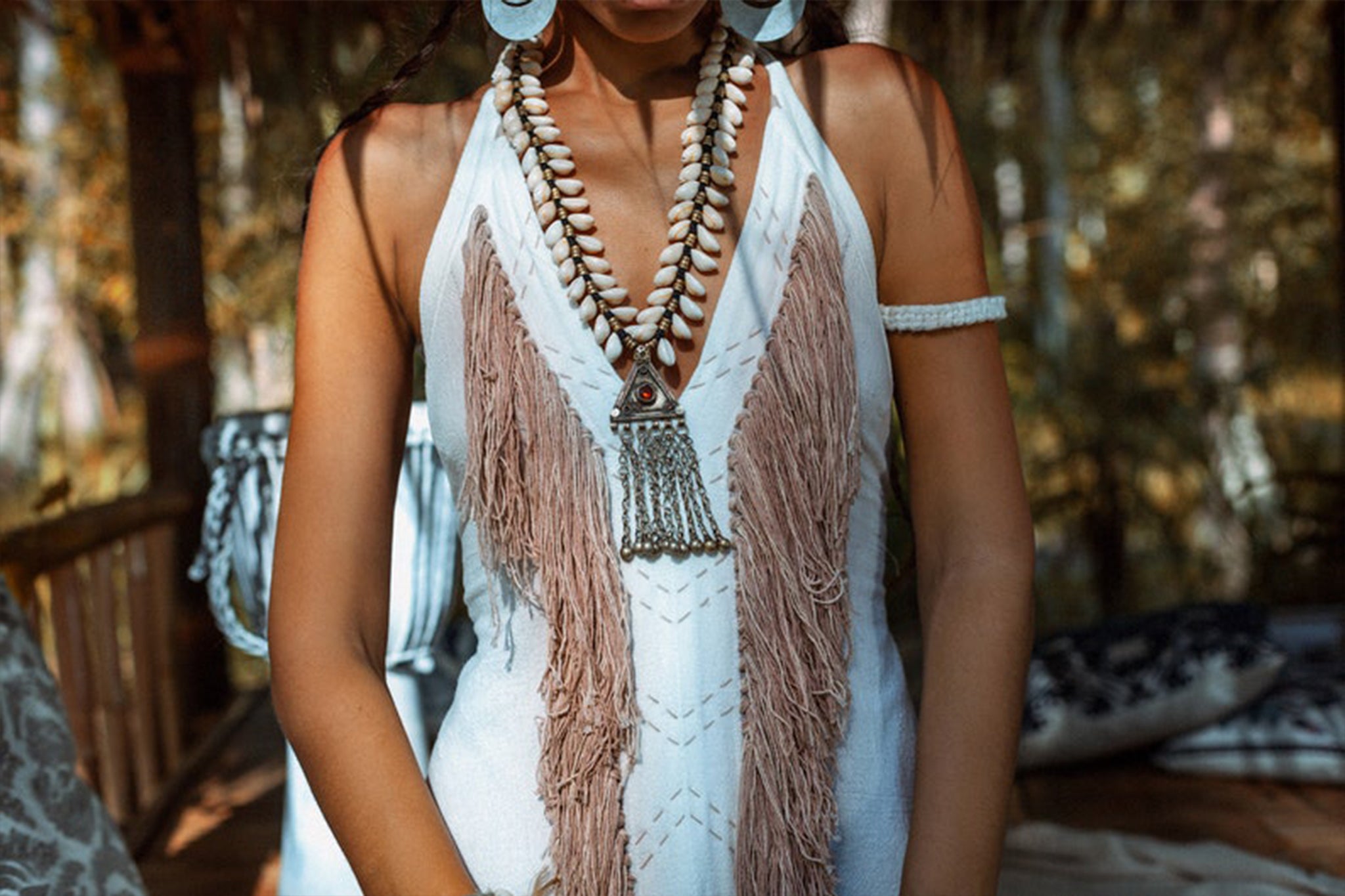 A woman dressed in the Off-White Boho Tribal Bohemian Dress, entirely handmade by AYA Sacred Wear, stands in a wooden structure. She accessorizes with layered shell necklaces and large white earrings complementing her braided hairstyle. The dress features organic fabric with fringe details and hand-made embroidery accents. The softly blurred background hints at a natural setting.