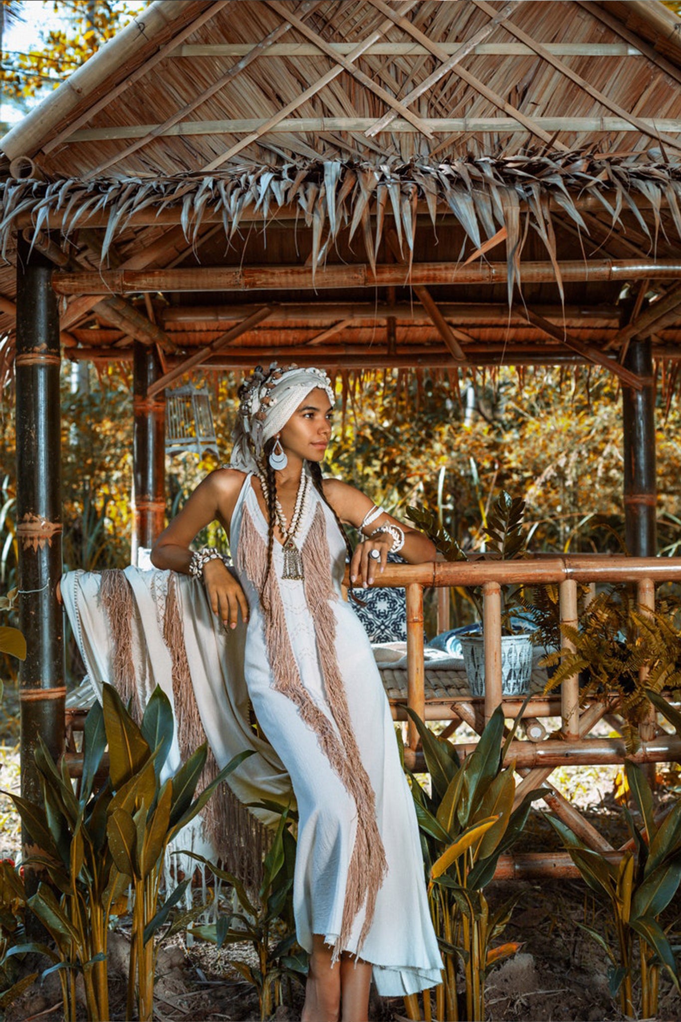 A woman in an Off-White Boho Tribal Bohemian Dress, entirely handmade by AYA Sacred Wear, leans against a bamboo railing in a rustic wooden hut. She pairs the dress with a headscarf and chunky jewelry, surrounded by tropical plants with golden foliage in the background.