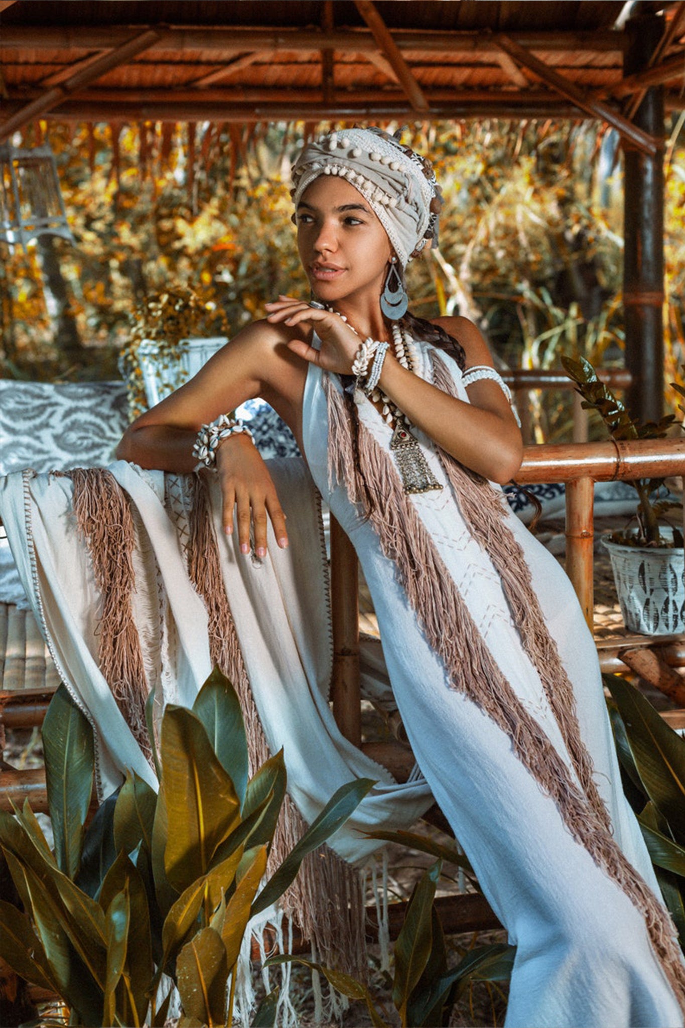 A woman wearing the Off-White Boho Tribal Bohemian Dress Entirely Handmade by AYA Sacred Wear and a headwrap poses in a tropical setting. She is adorned with layered necklaces and bracelets, leaning against a wooden structure surrounded by decorative plants. The handmade embroidery amplifies the scene's bohemian and natural vibe.