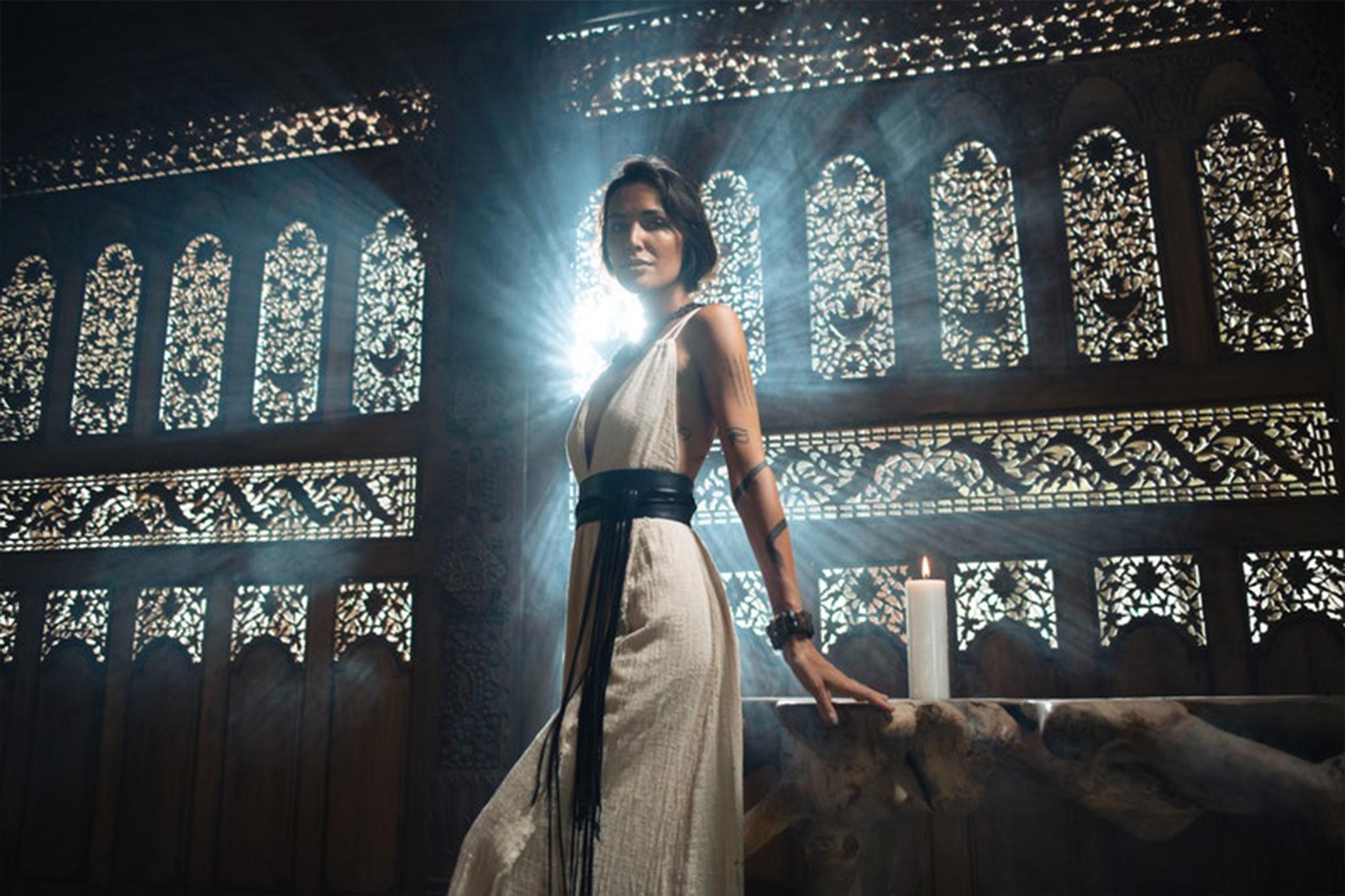 An elegant woman wearing an Off-White Boho Wedding Dress with Open Back by AYA Sacred Wear stands beside a table, her figure highlighted by the light streaming through ornate windows. She is in a dimly lit room adorned with intricate designs on the back wall, adding to the dramatic and mystical atmosphere.