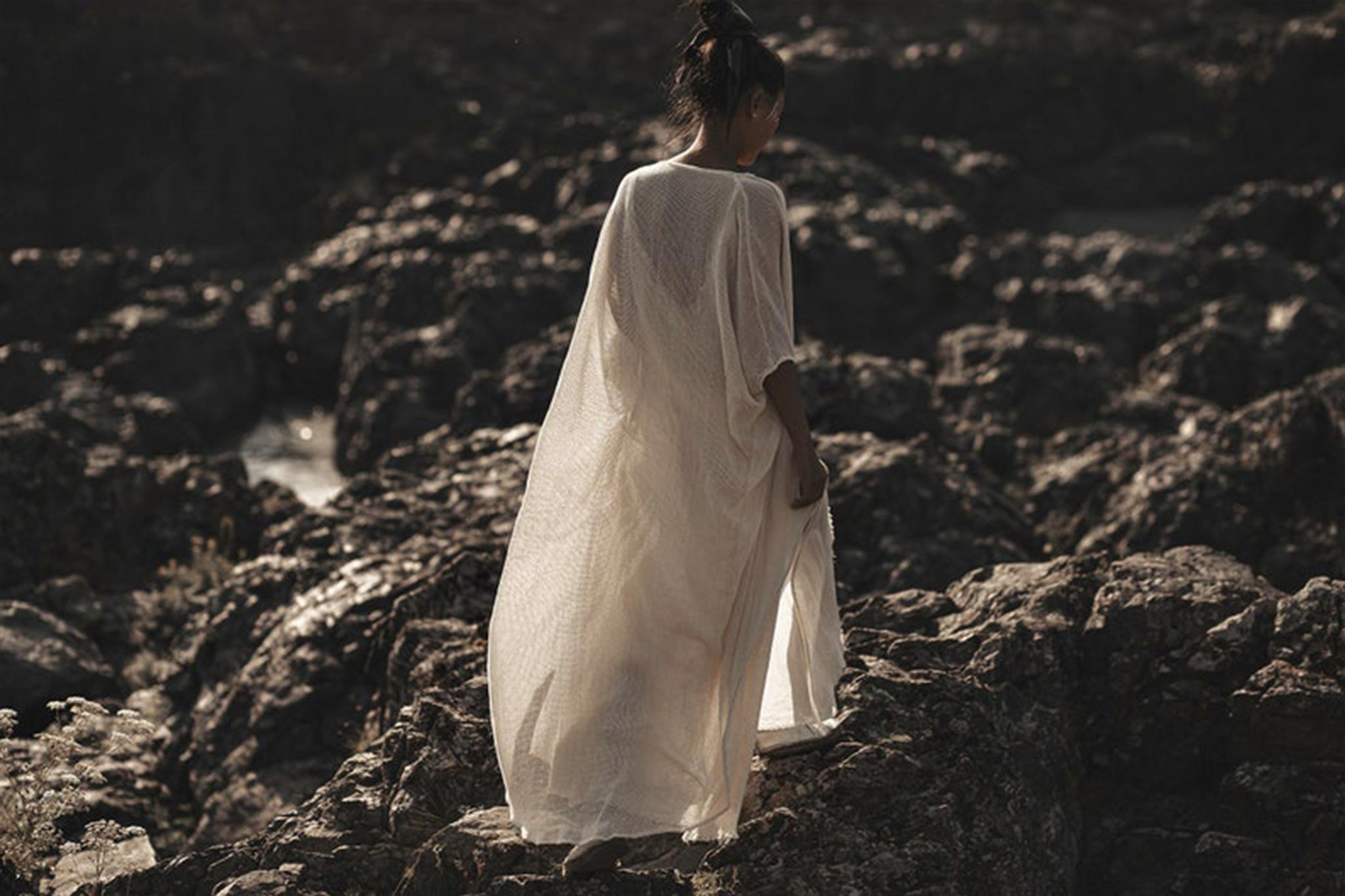An individual draped in an Off-White Cotton Boho Kimono Cover-up by AYA Sacred Wear stands gracefully on rocky terrain. This exquisite piece, functioning as a bridal cape or poncho robe, enhances the serene and mysterious atmosphere with its sustainable fashion flair against a backdrop of rugged, dark rocks.
