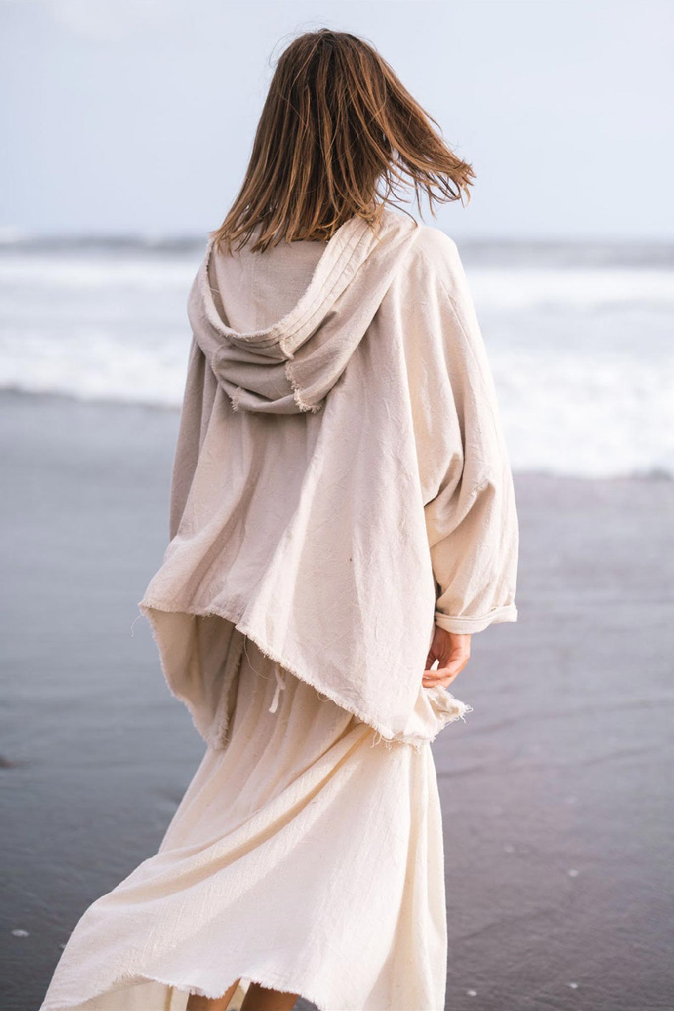 Wearing an Off-White Cotton Warm Jacket with Hood in Bohemian Style by AYA Sacred Wear, a person stands on the beach, facing the ocean. Their hair gently sways in the wind as waves crash in the background under a cloudy sky.