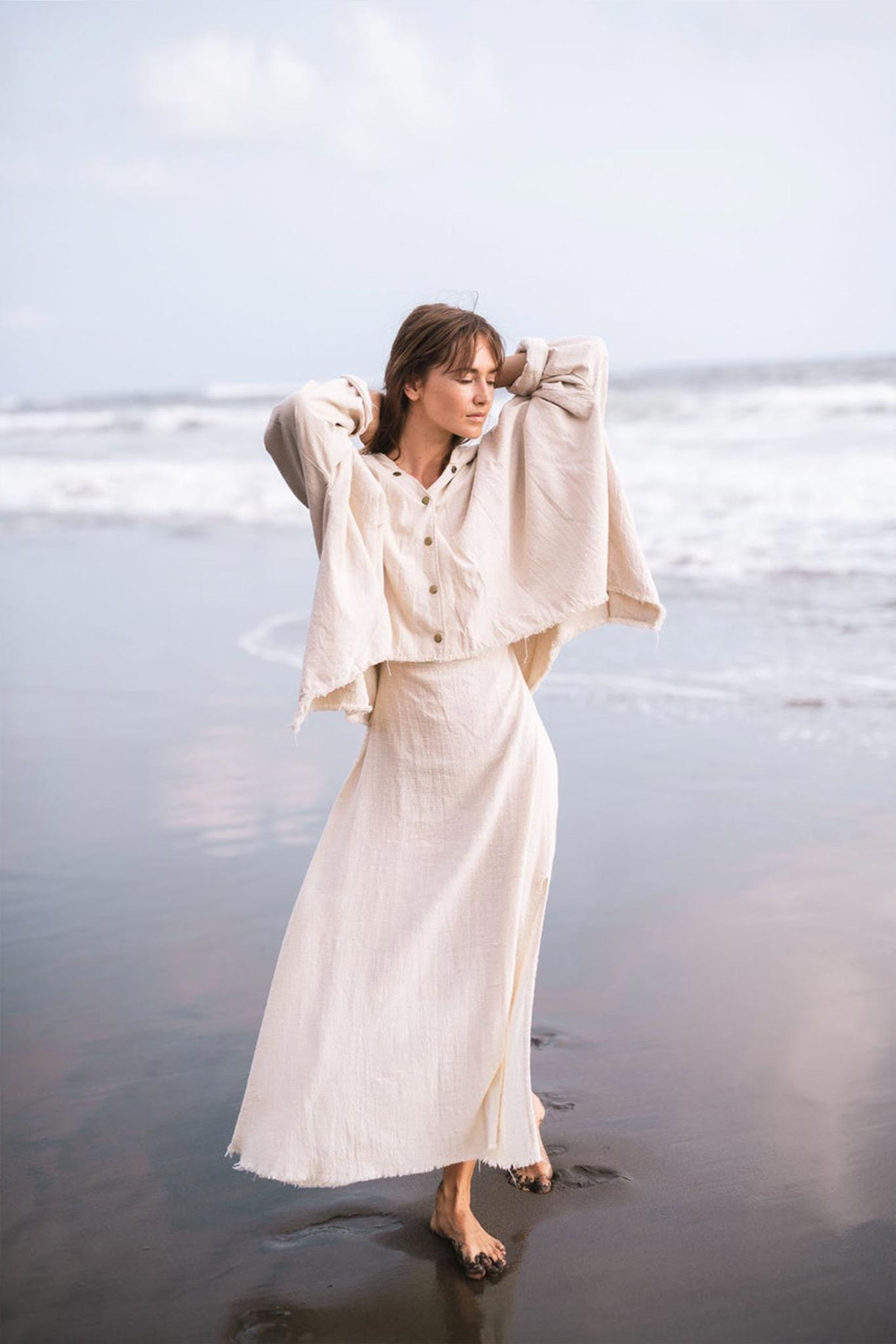 A woman stands on a beach with waves in the background, wearing the Off-White Cotton Warm Jacket with Hood from AYA Sacred Wear. Her arms are raised, and she gazes thoughtfully into the distance. The scene is serene and minimalist.