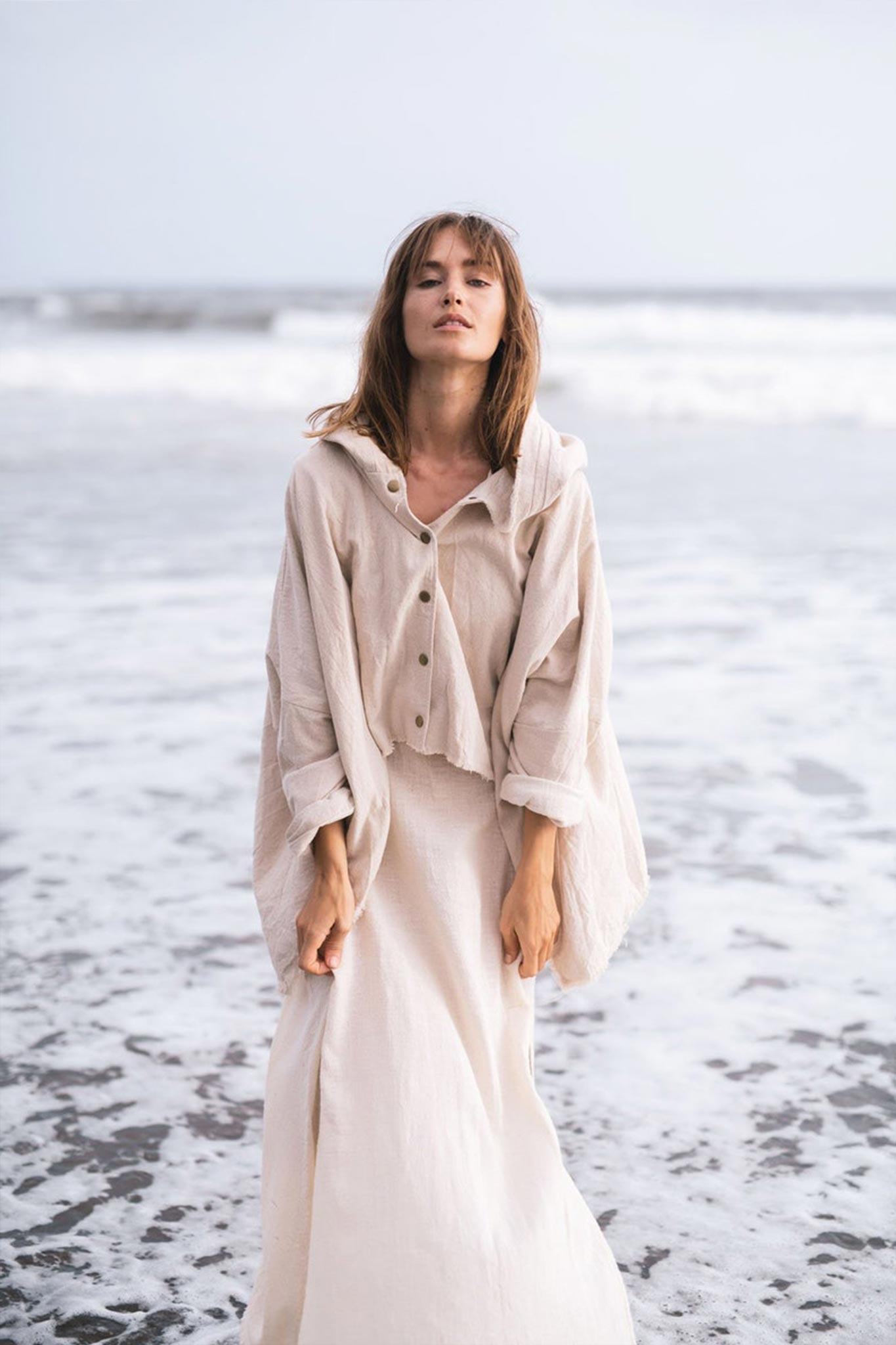 On a tranquil shoreline, a person is enveloped in an Off-White Cotton Warm Jacket with Hood by AYA Sacred Wear, exuding Bohemian style. As ocean waves gently kiss their feet, sea foam swirls across the sand, while the overcast sky adds a peaceful and serene ambiance to the scene.