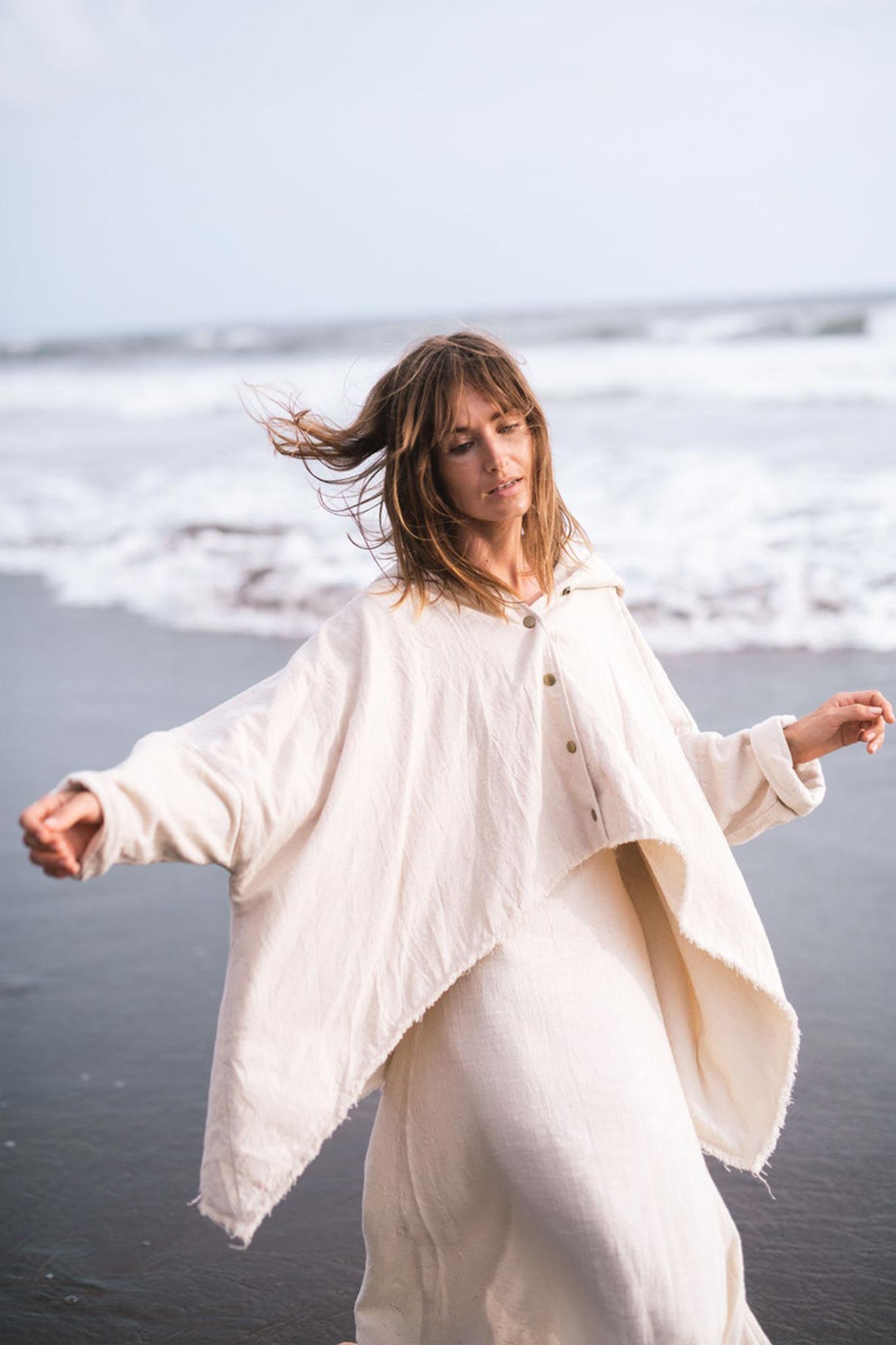 A woman with long hair stands on a beach, wrapped in the Off-White Cotton Warm Jacket with Hood Bohemian Style by AYA Sacred Wear, her arms gently outstretched as the ocean waves and cloudy sky create a serene backdrop.