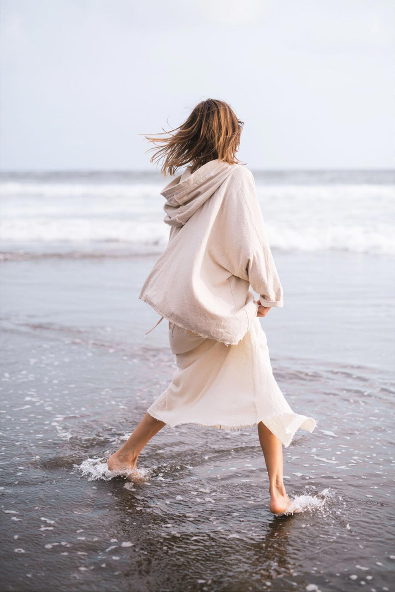 Dressed in an Off-White Cotton Warm Jacket with Hood Bohemian Style from AYA Sacred Wear, an individual strolls barefoot along the shoreline. The ocean waves and wet sand are visible as they face away from the camera, their hair and naturally dyed attire gently tousled by the breeze.