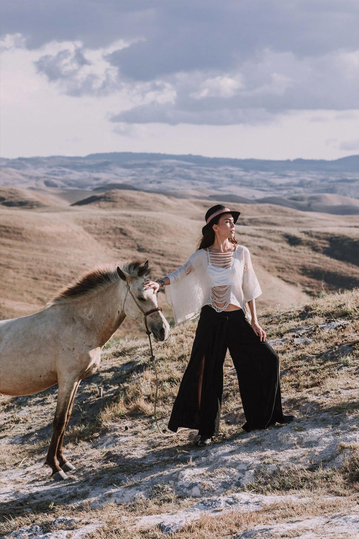 Dressed in an Off-White Hand Loomed Raw Cotton Top by AYA Sacred Wear and black wide-leg pants, a person stands on a hilly grassland, holding the reins of a brown horse. With a hat atop their head, they gaze into the distance under a cloudy sky. Hills extend across the landscape in the background, creating an evocative scene.