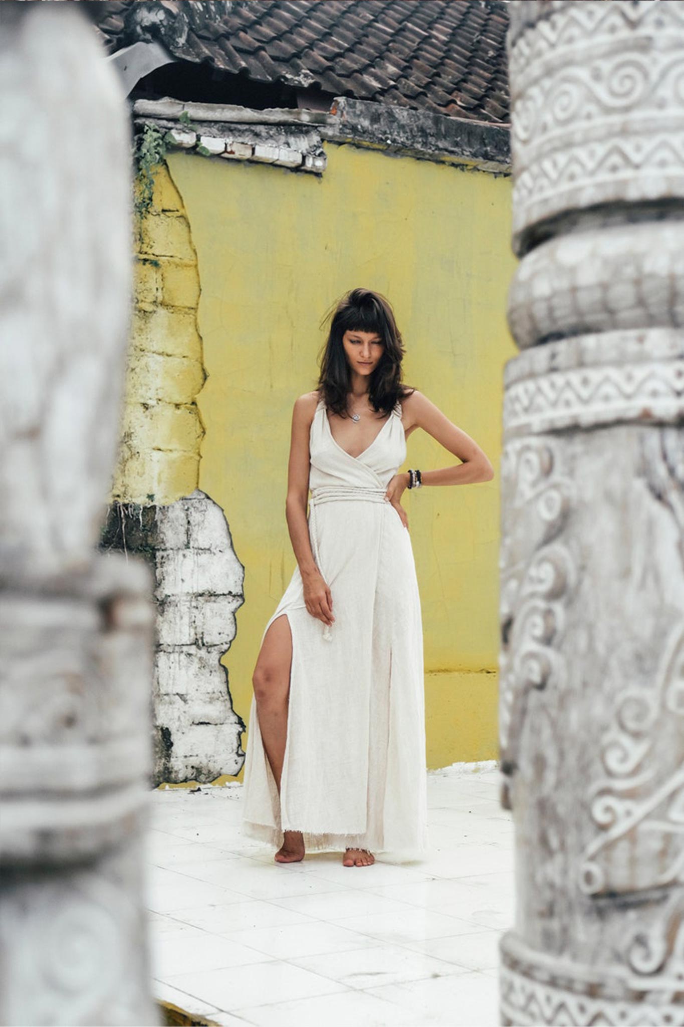A woman is standing barefoot on a white tile floor, dressed in an Off-White Handwoven Wrap Maxi Dress with a Hand Loomed Back by AYA Sacred Wear. She is framed by intricately carved stone pillars. Behind her, a yellow wall with peeling paint beneath a brown tiled roof forms a serene backdrop for her contemplative expression.