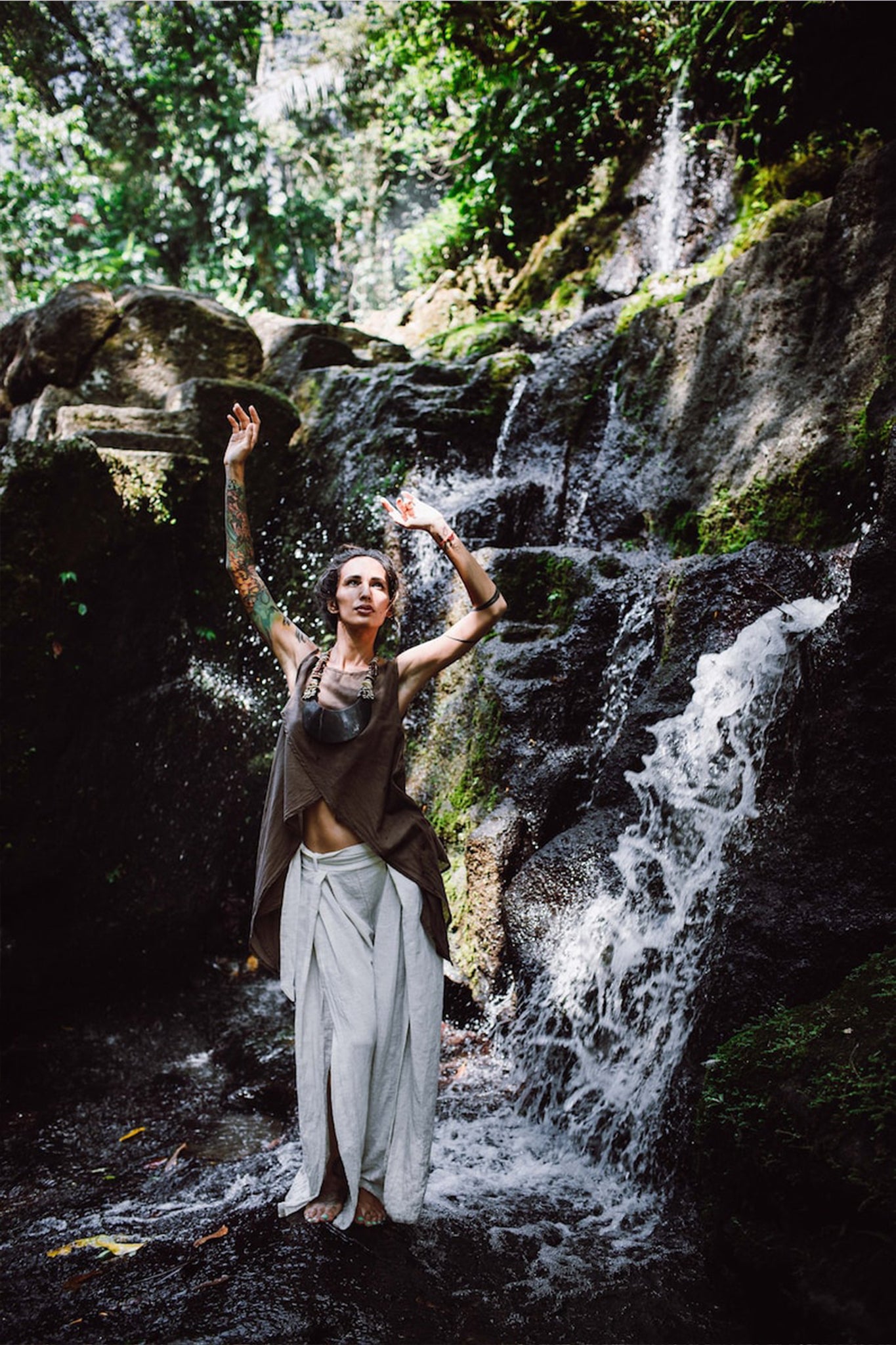 Standing barefoot beside a cascading waterfall, a person wears Off-White Linen Boho Pants with High Slit from AYA Sacred Wear along with a sleeveless top. Their arms are lifted upwards amidst lush greenery and sunlight streaming through the trees.